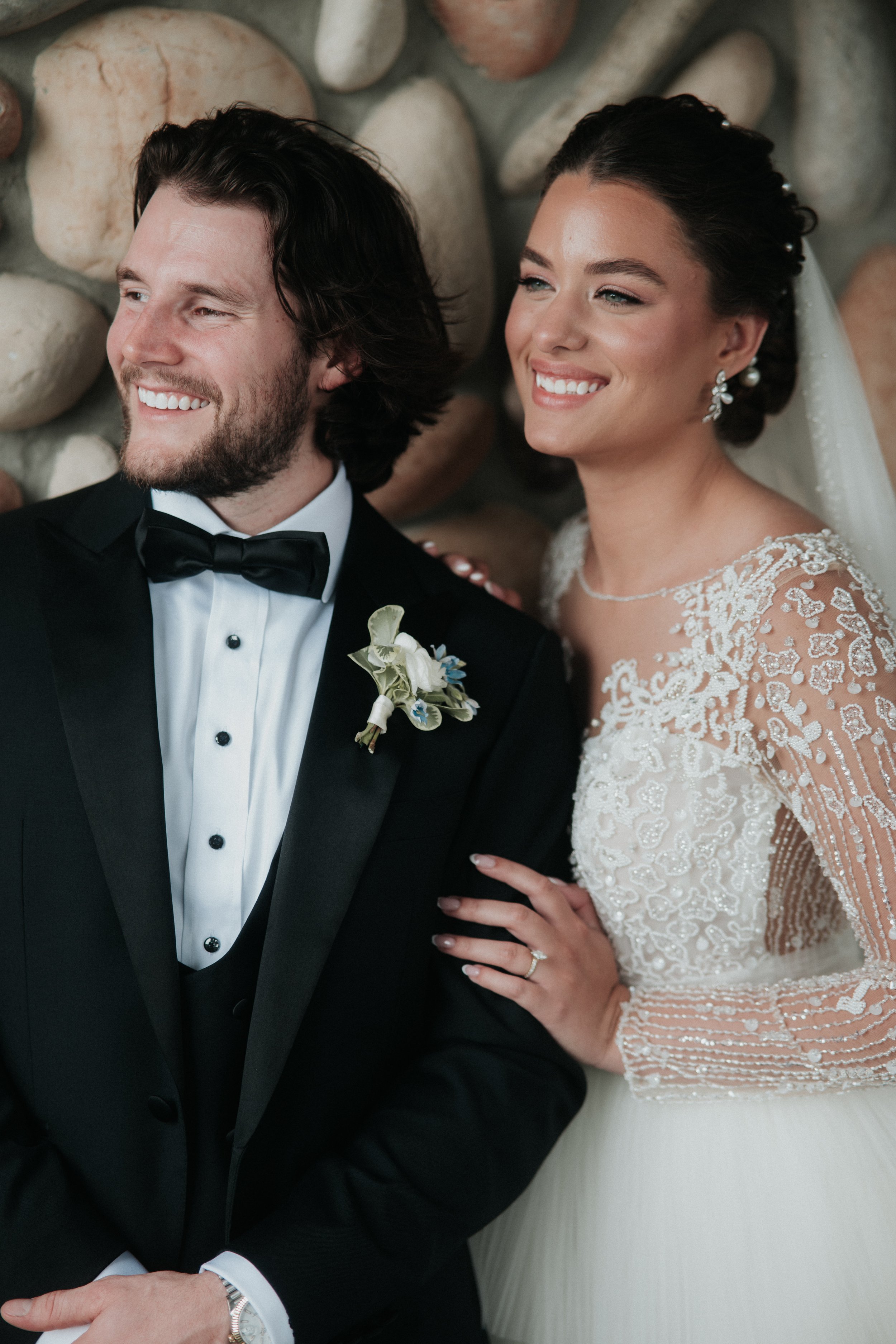 A bride and groom dressed in wedding attire, smiling and looking to the side, against a background of large stones.