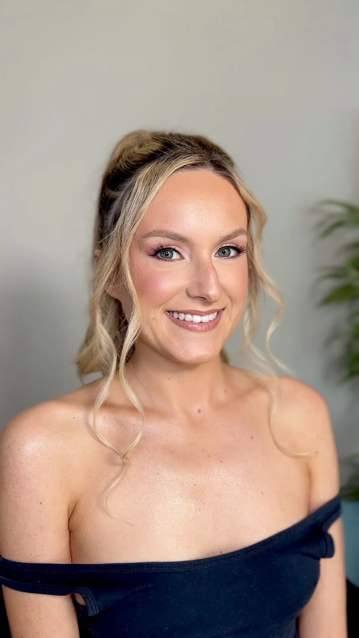 A young woman with blonde hair styled in loose curls, wearing a black off-the-shoulder top, smiling at the camera, with a blurred green plant in the background.