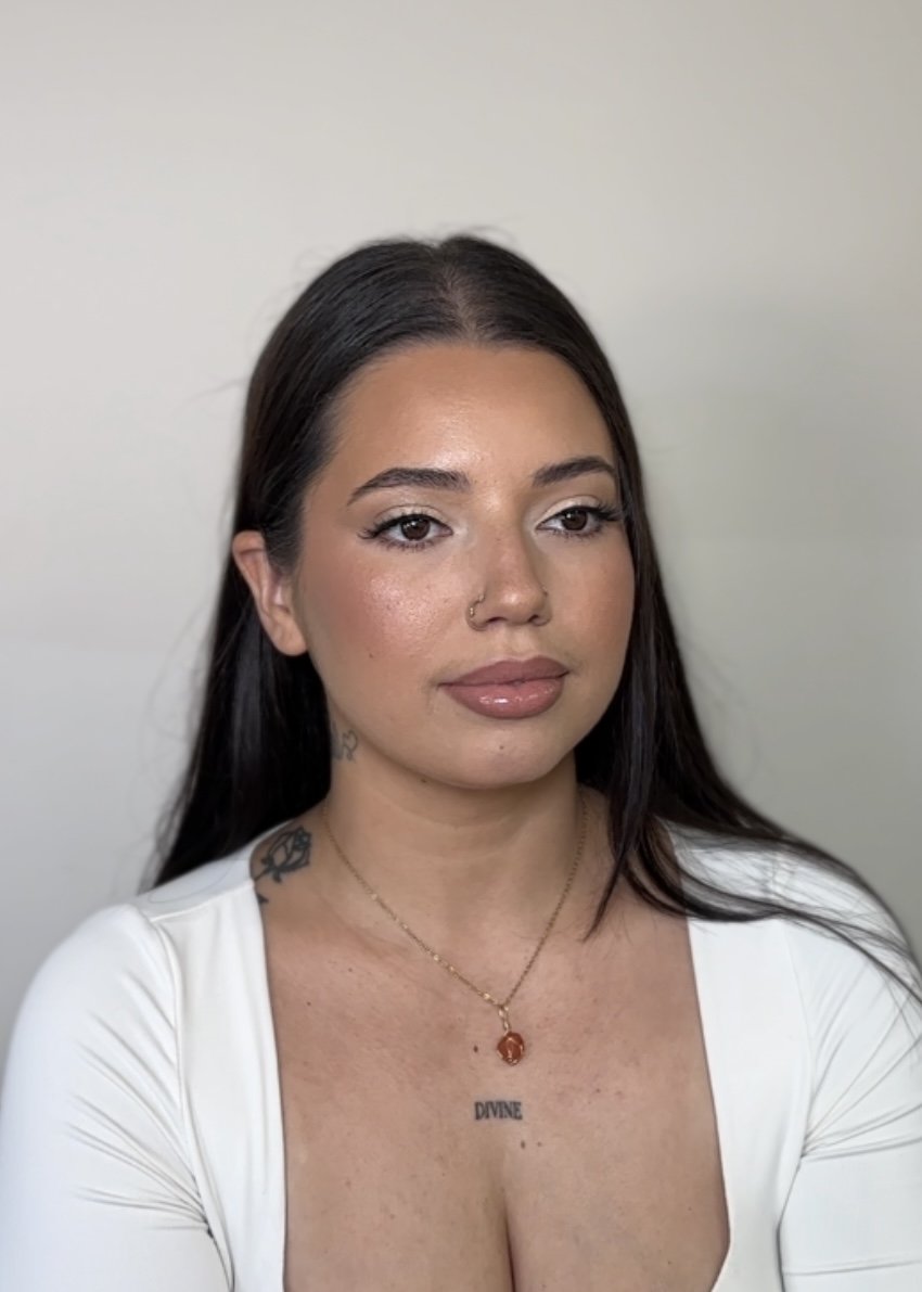 A woman with long dark hair and light skin, wearing a white top and a necklace with a pendant, standing indoors against a plain light-colored wall.