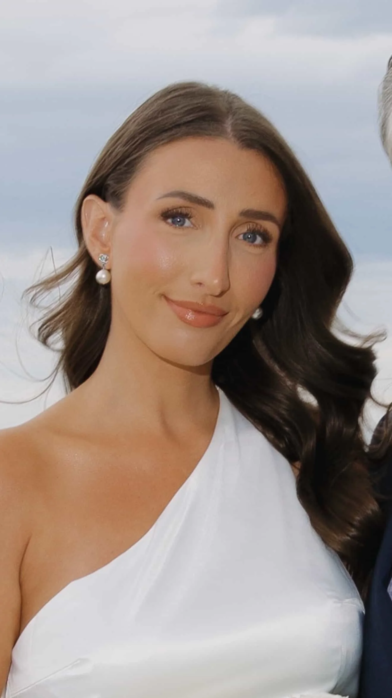 A woman with brown hair, blue eyes, and pearl earrings, wearing white clothing, smiling at the camera outdoors with a cloudy sky background.