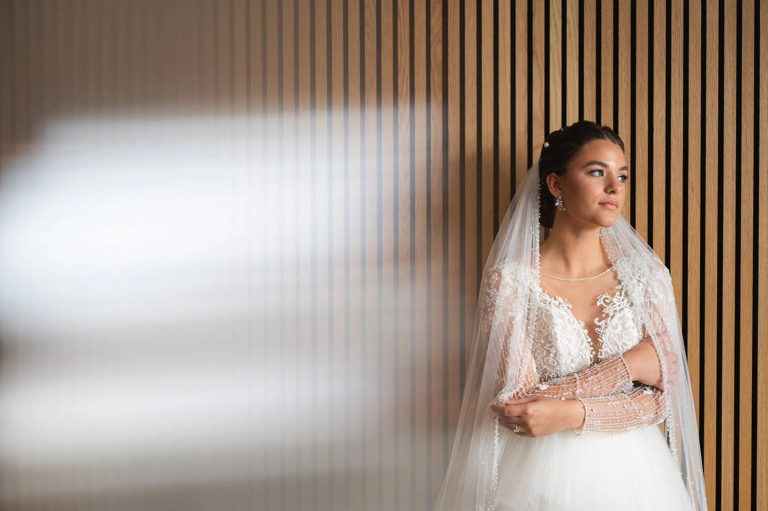 Bride in wedding dress and veil standing against a wooden panel wall, looking to the side.