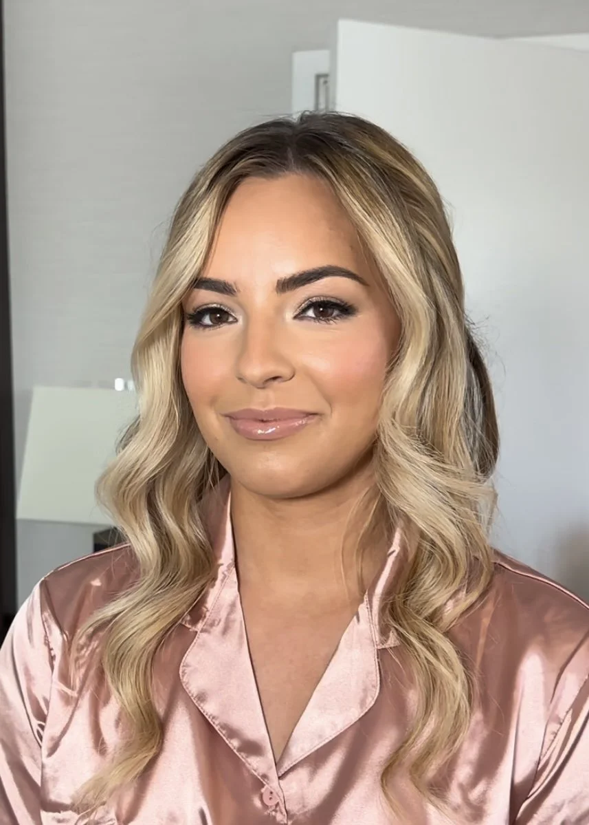 Close-up of a woman with long wavy blonde hair, wearing a pink satin pajama top, smiling softly inside a modern room.