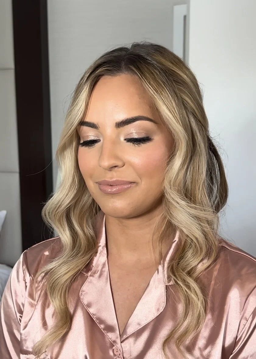 A woman with styled blonde hair and makeup, wearing pink satin pajamas, indoors with a neutral background.