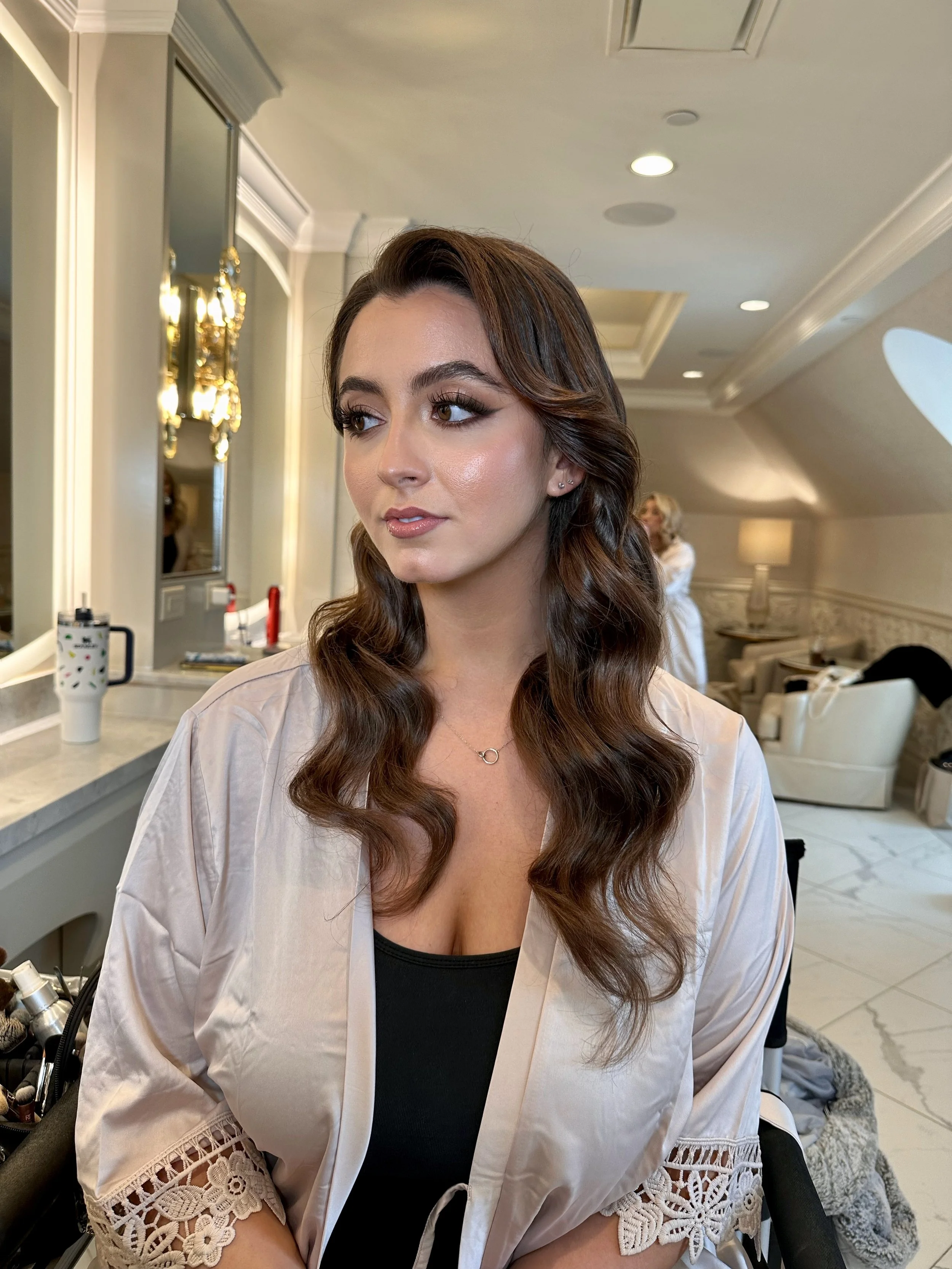 A woman with long, wavy brown hair and makeup, wearing a satin robe with lace sleeves, sitting in a room with elegant decor.