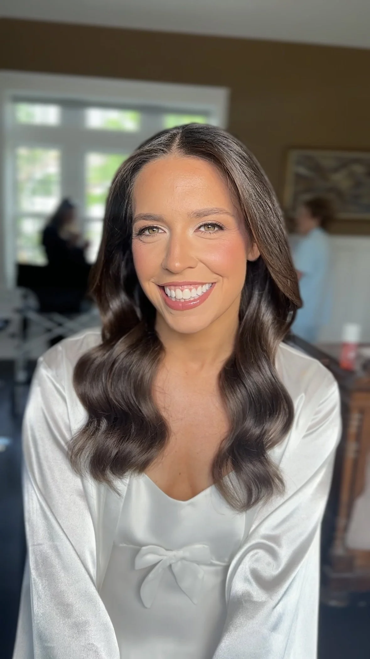 A woman with long wavy brown hair and light skin smiling at the camera in a well-lit room.