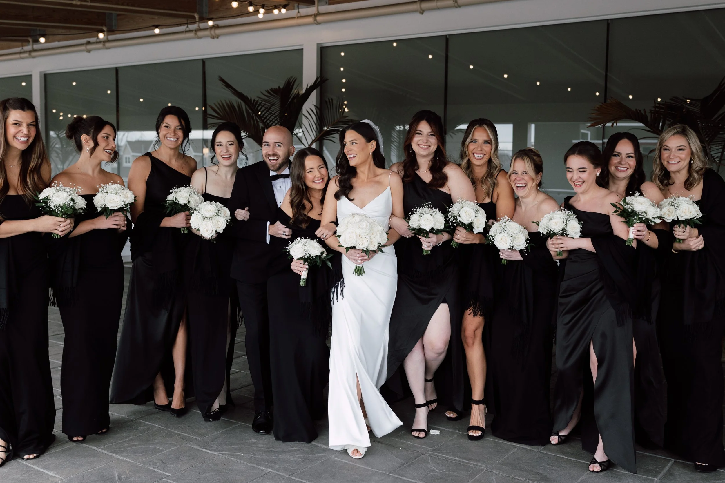 Wedding party with the bride in white and bridesmaids in black dresses holding white bouquets, standing outside under a covered area with large windows and palm plants.