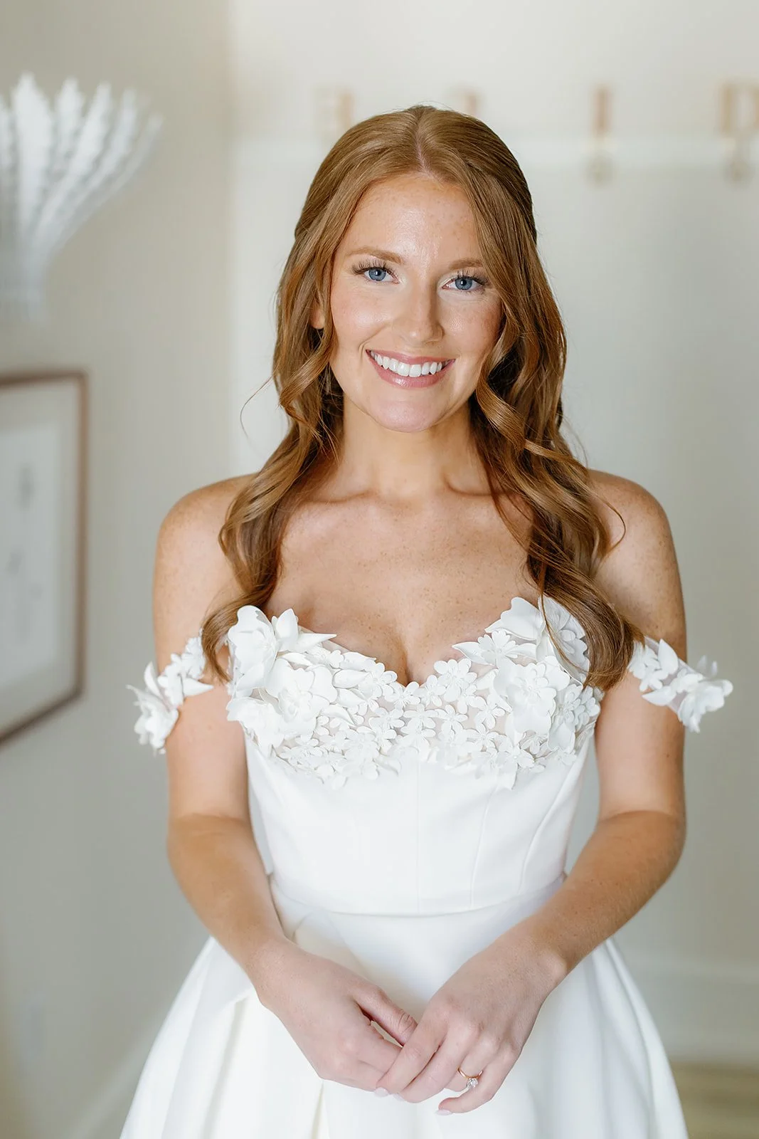 A woman with long, wavy auburn hair and blue eyes, smiling in a white wedding dress with floral lace details on the off-the-shoulder neckline, standing indoors with a light-colored wall and framed pictures in the background.