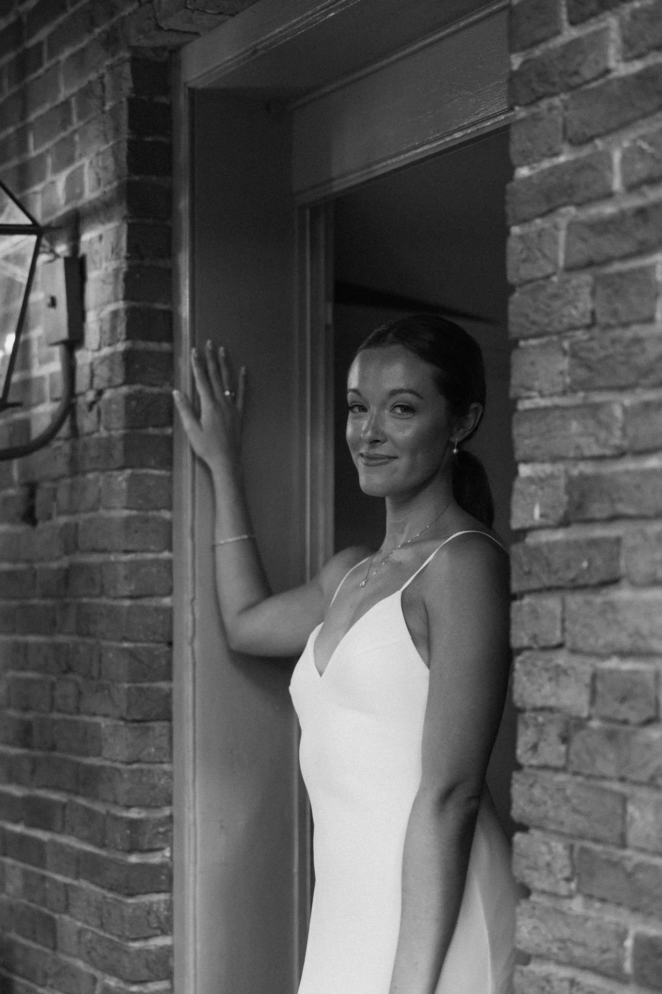 A woman in a white dress standing at a doorframe, smiling, with her hand resting on the wall. The photo is in black and white, with brick walls on either side.