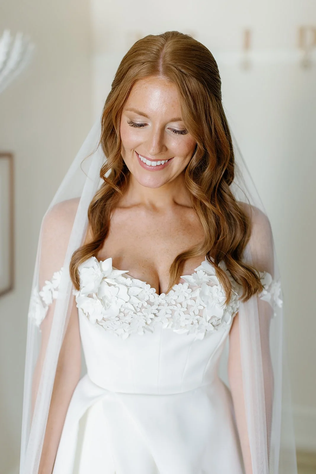 A bride in a white wedding dress with floral appliqué, smiling and looking down, with long wavy auburn hair and a veil.