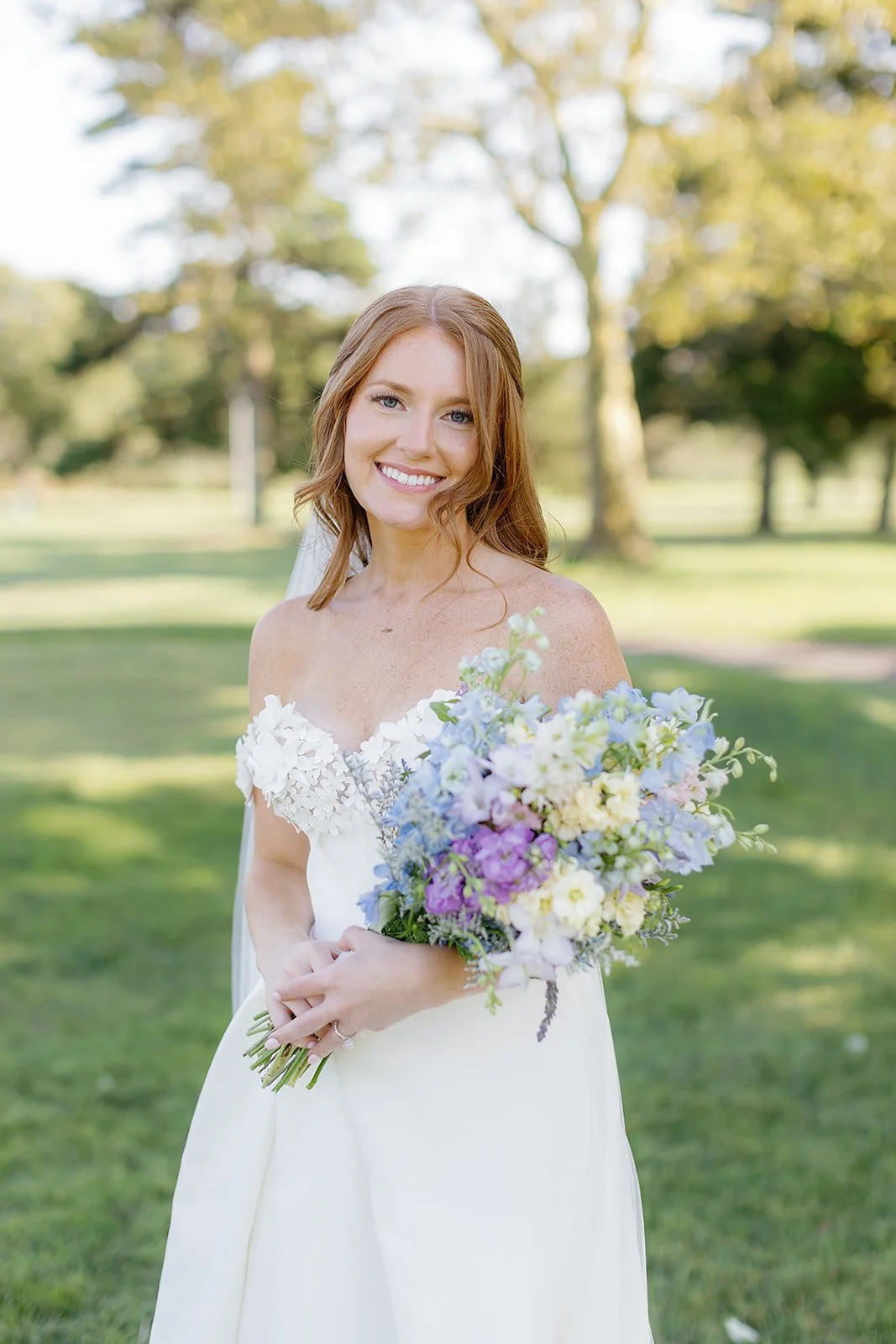 A smiling bride holding a colorful bouquet of flowers outdoors on a sunny day.