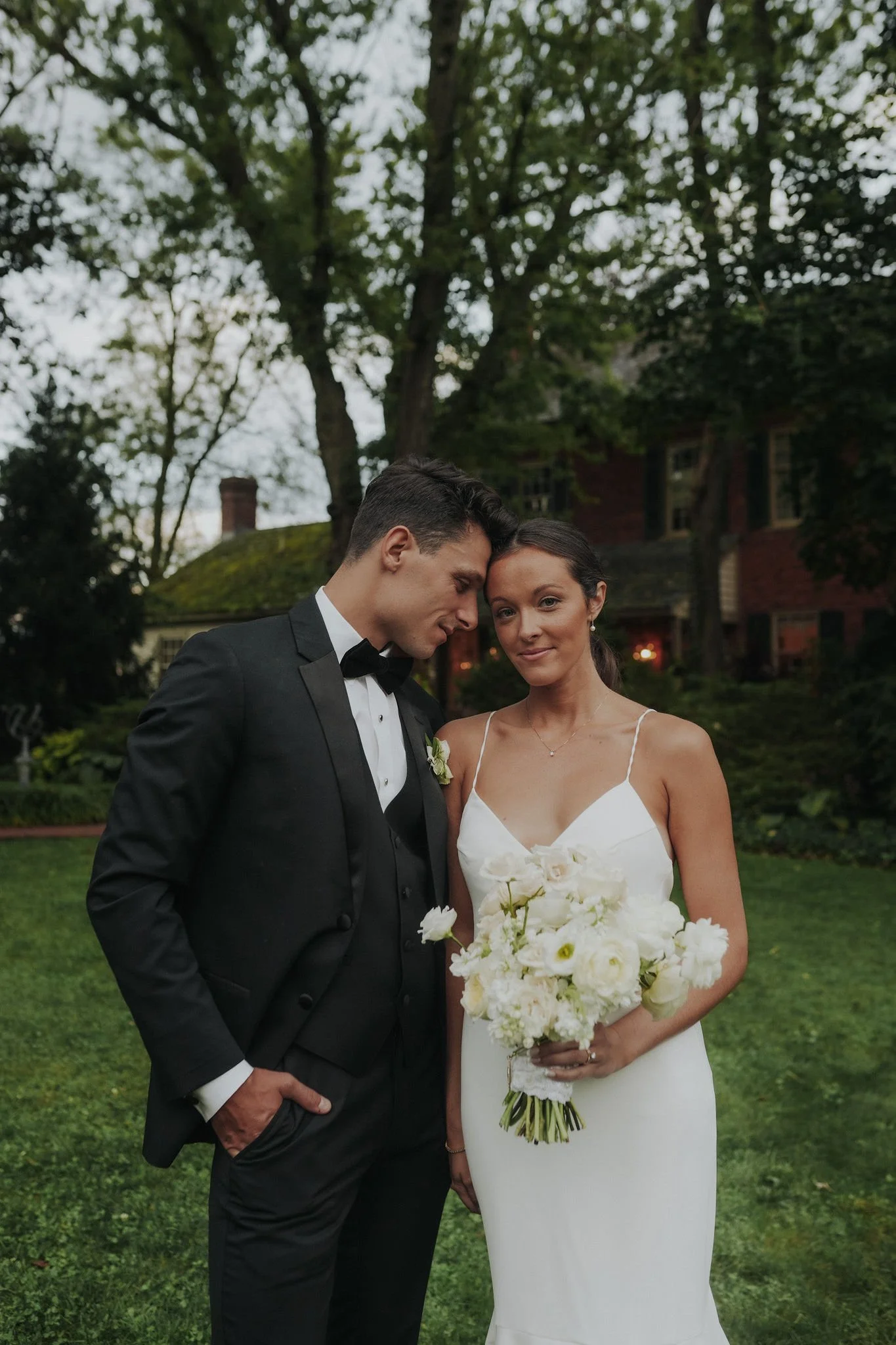A newlywed couple standing outdoors in a garden at dusk, with the groom in a tuxedo and the bride in a white wedding dress holding a bouquet of white flowers.