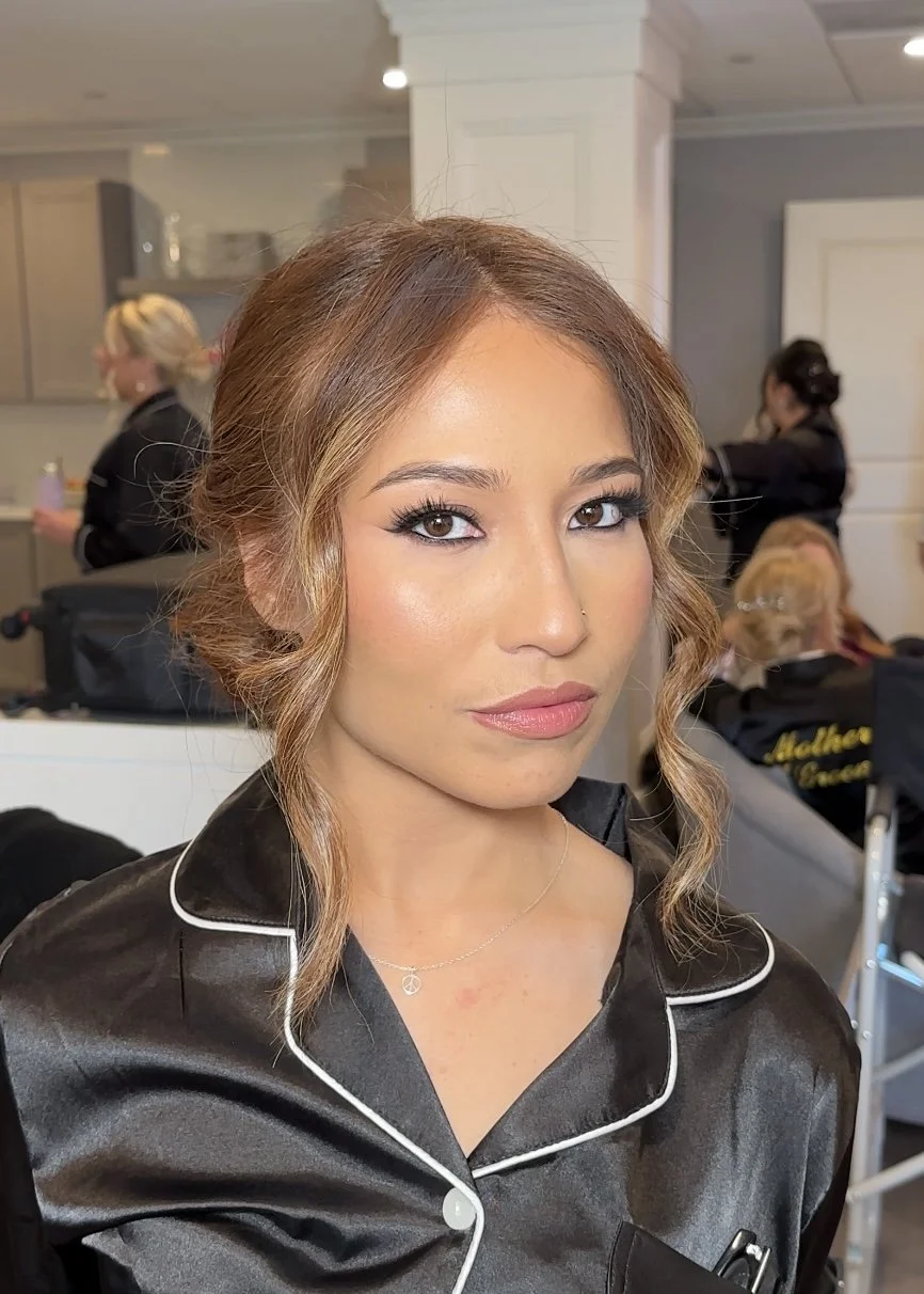 A woman with wavy light brown hair and makeup, wearing black satin pajamas with white piping, looking at the camera, in a room with people and makeup stations in the background.