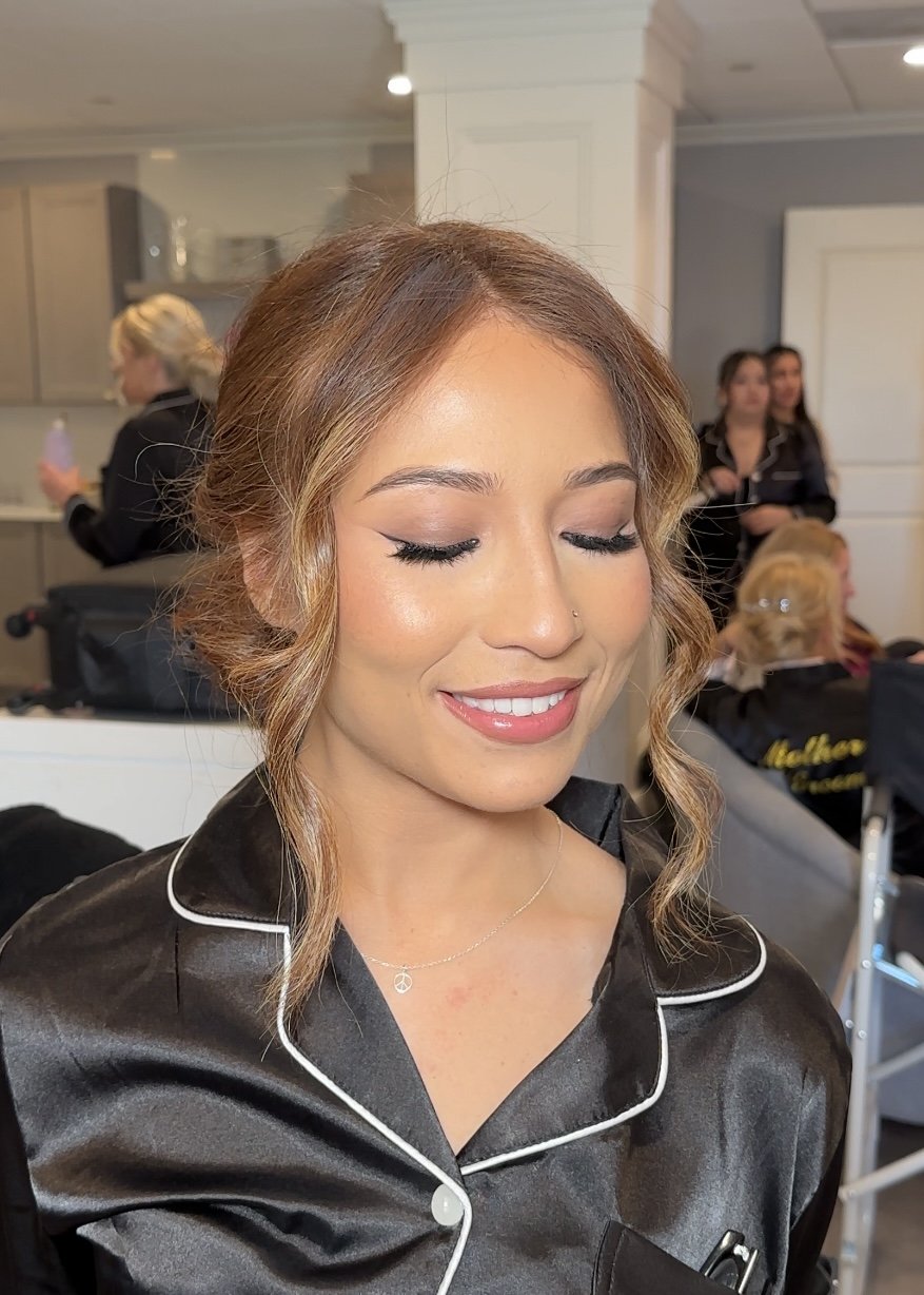 A young woman with wavy, light brown hair and closed eyes, wearing makeup, a black satin pajama top with white piping, and a delicate necklace, smiling softly in an indoor setting with a few people in the background.
