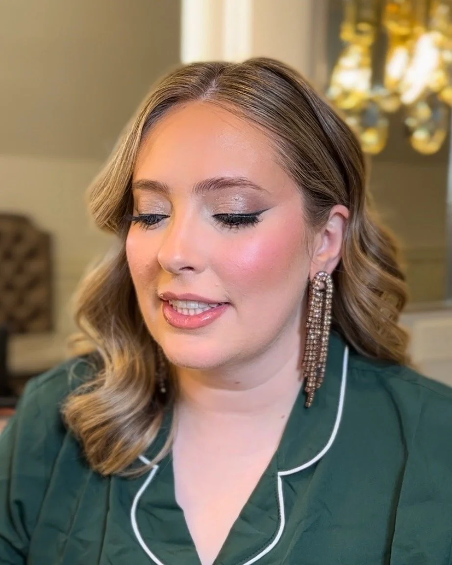 A woman with light brown, wavy hair and makeup, wearing large dangling earrings and a green top with white piping, looking downward indoors.