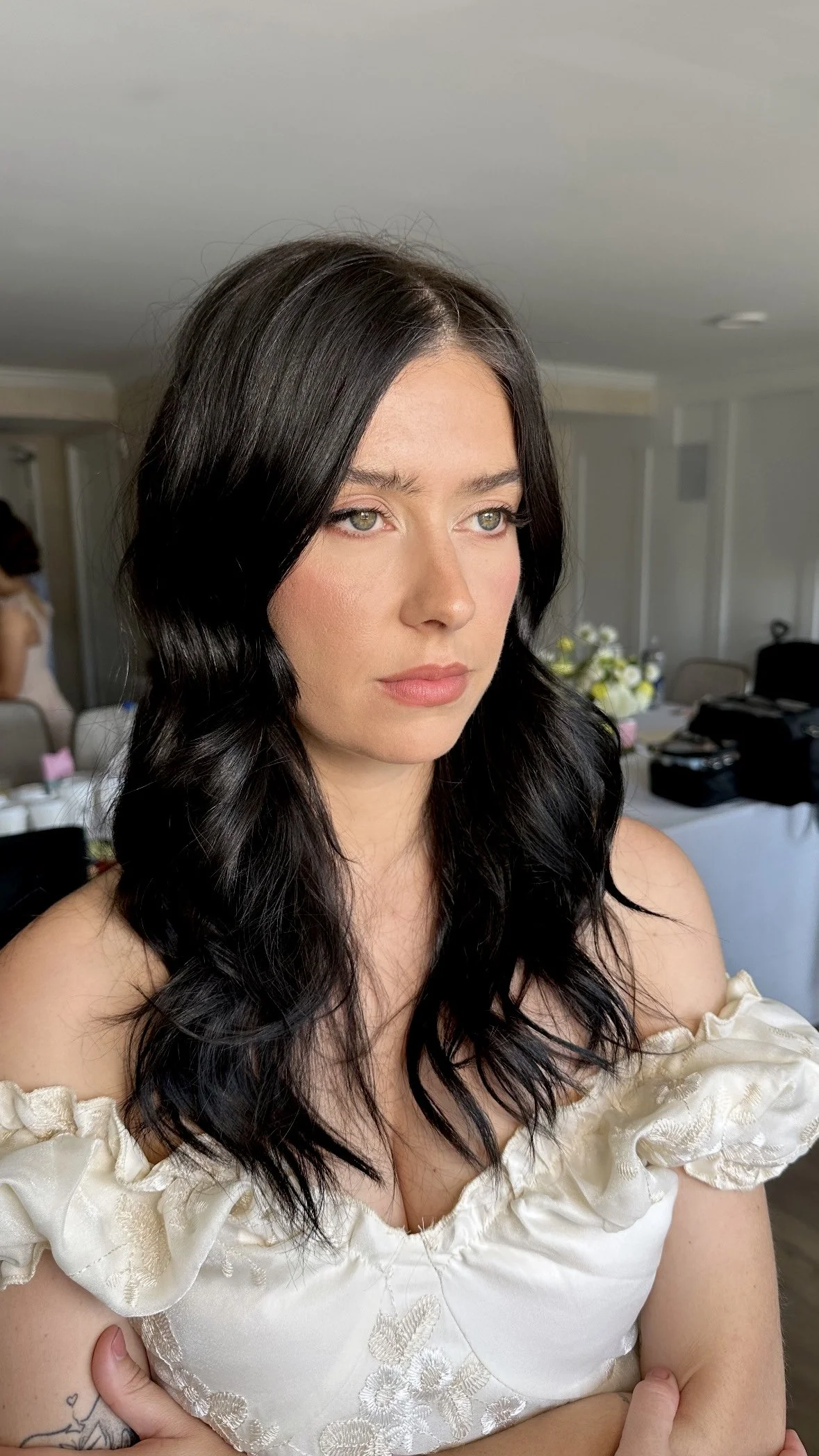 A woman with long black wavy hair and fair skin wearing a white off-shoulder top, standing indoors with a neutral expression.