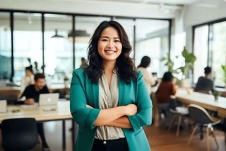 A asian woman with medium length black wavy hair stands in a creative environmet, with people in the background working on computers. She's wearing a green jacket, with her arms crossed, and looks confident.