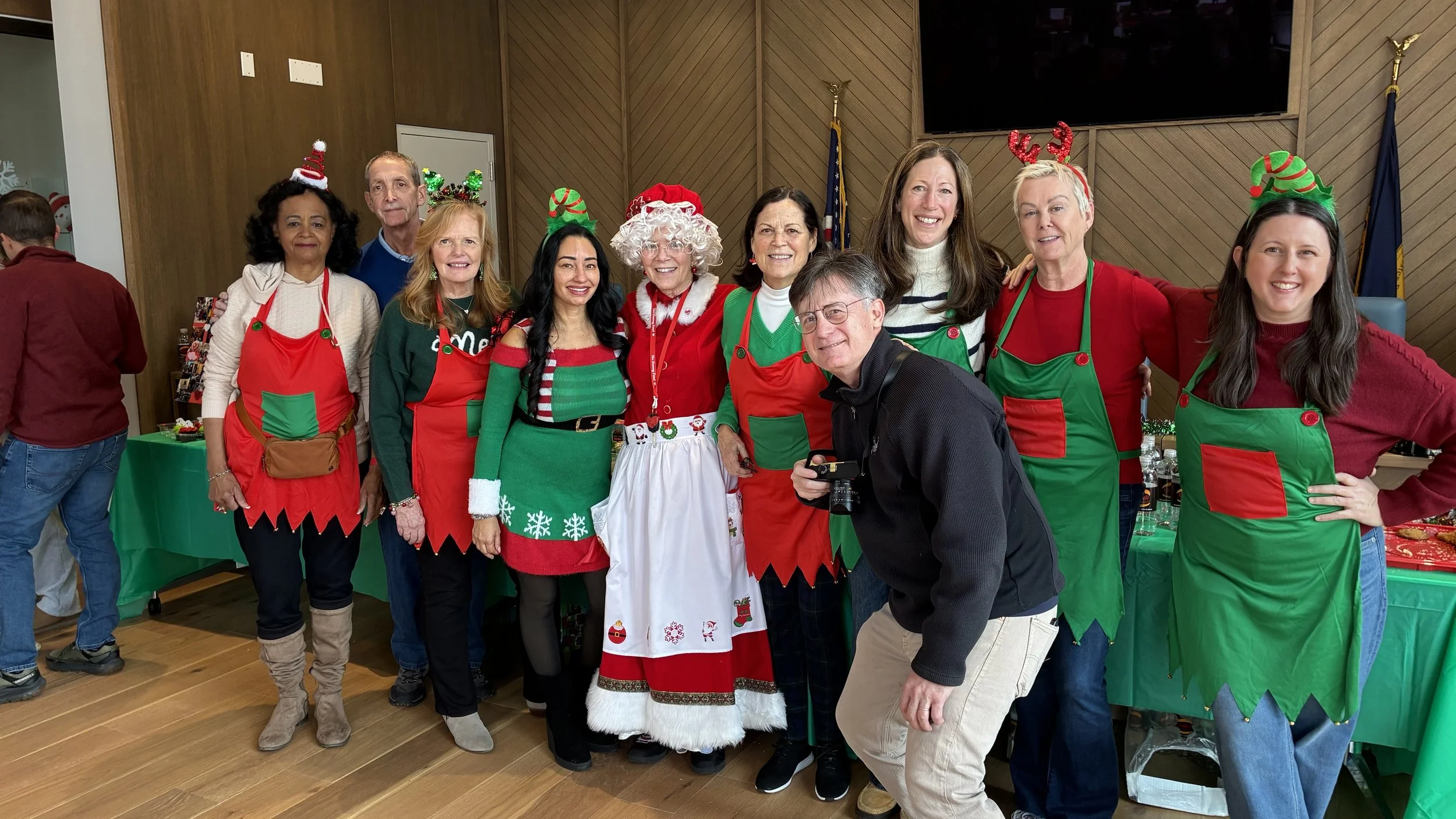 A group of people dressed in Christmas-themed clothing and accessories, posing together indoors with holiday decorations and tables in the background.