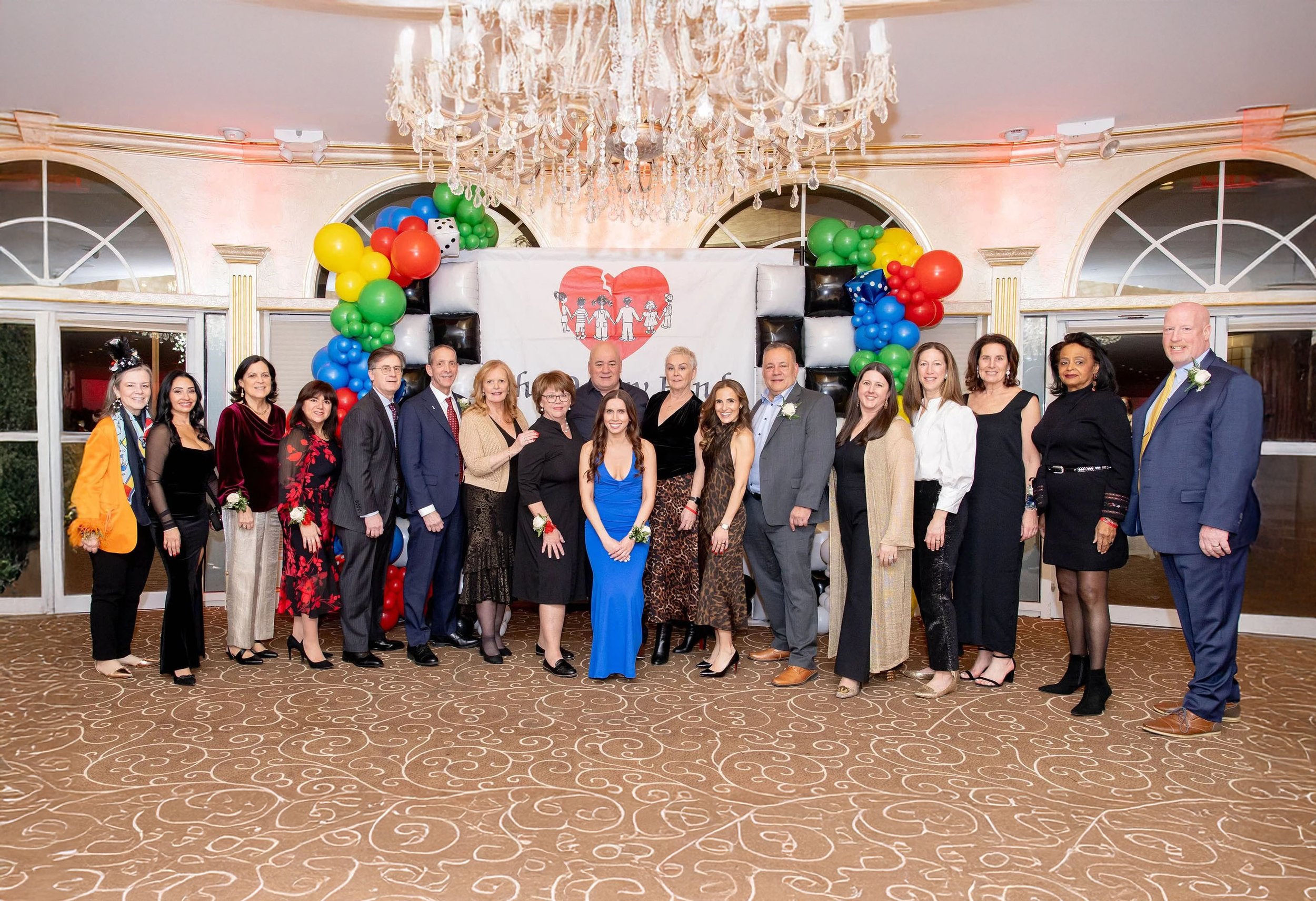 Group of people in formal attire standing together at a celebration, with a colorful balloon arch and a cake as backdrop, in a decorated banquet hall with a chandelier and patterned carpet.