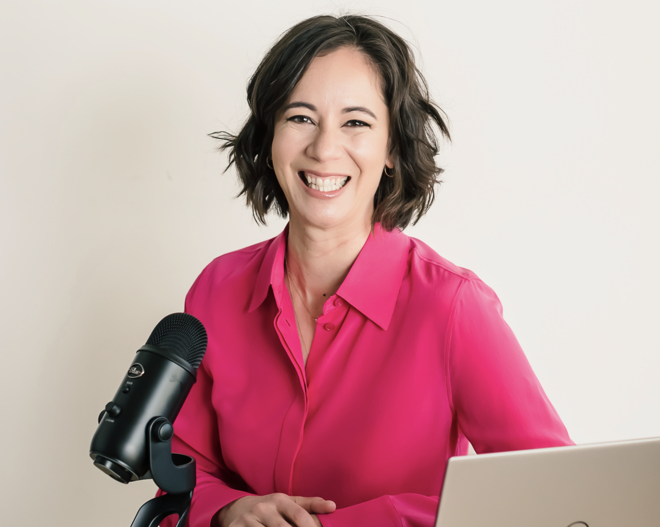 A woman with short, dark hair and wearing a bright pink blouse, smiling and sitting at a desk with a microphone and a laptop in front of her.