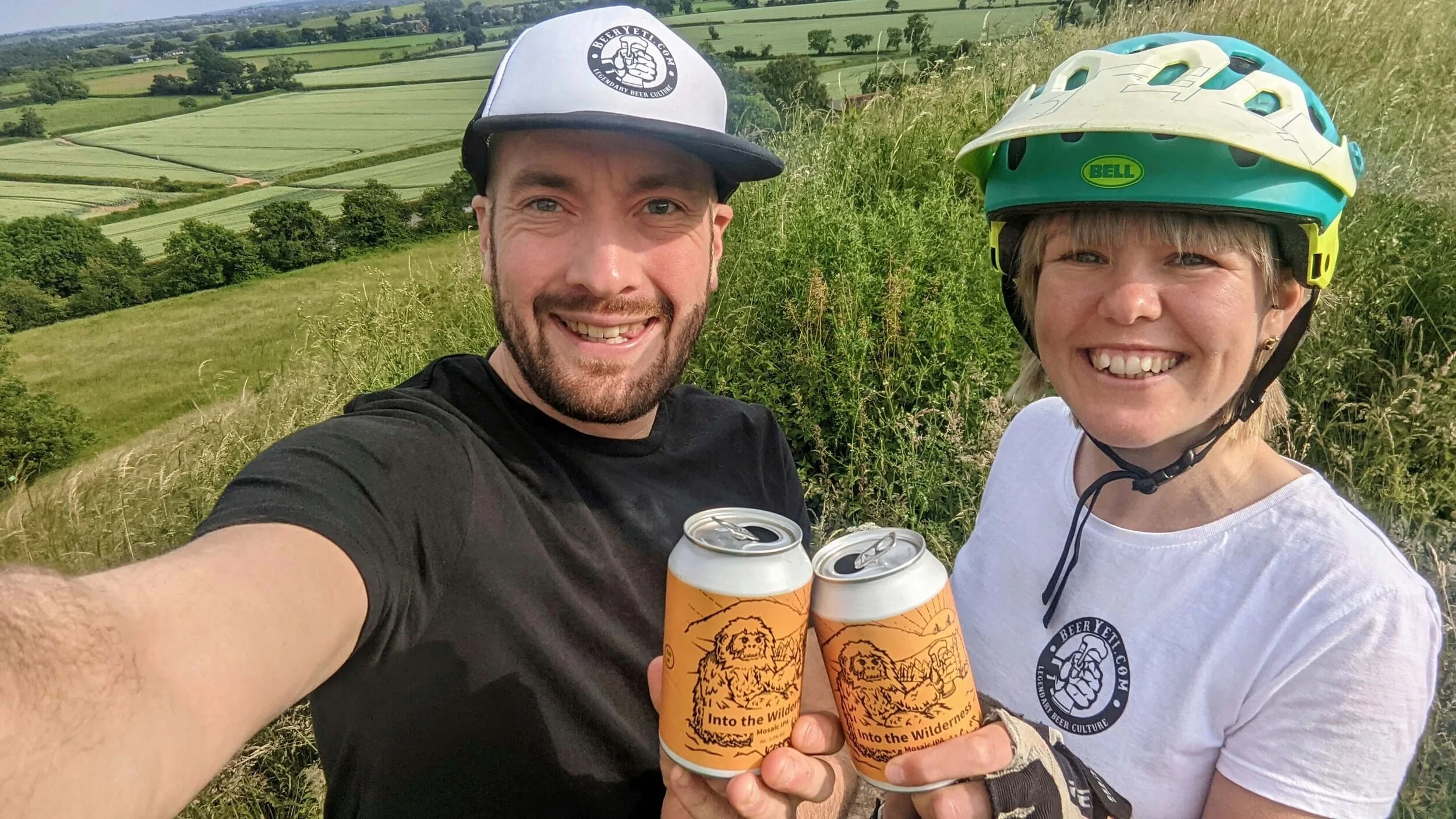 A picture of Tim & Lisa Jones from BeerYeti holding up their new cans of beer on a bike ride with a beautiful countryside backdrop