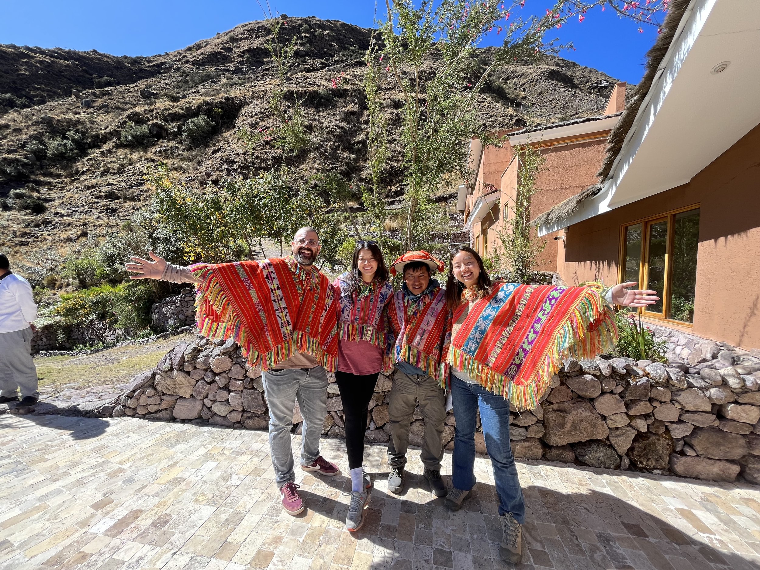 A group of four people wearing colorful traditional clothing, standing outdoors with a mountainous landscape in the background. They are smiling and have their arms outstretched, with a building on the right side of the image.