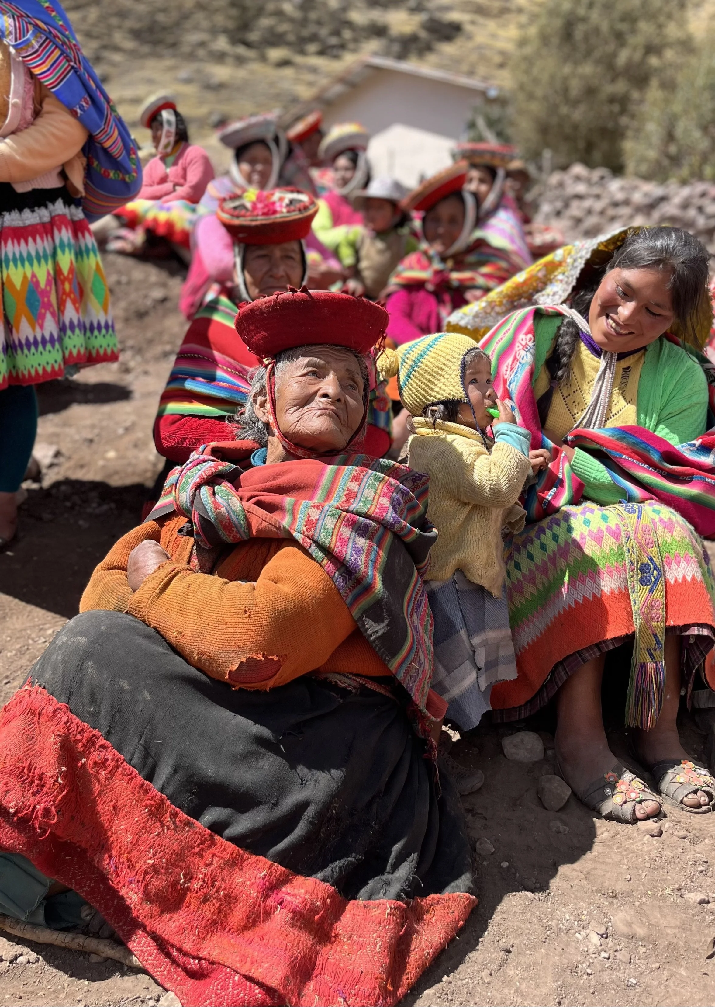 Group of indigenous women and children wearing colorful traditional clothing and hats, sitting outdoors on dirt ground, with some smiling and others looking serious, in a rural setting.