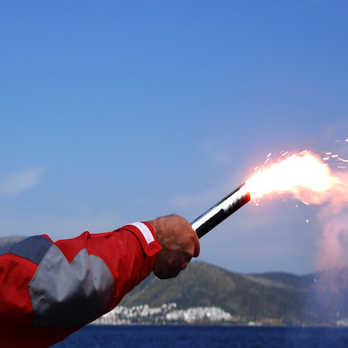 A man's arm in a red waterproof jacket holding a lit flare demonstrating Safety & Sea Survival