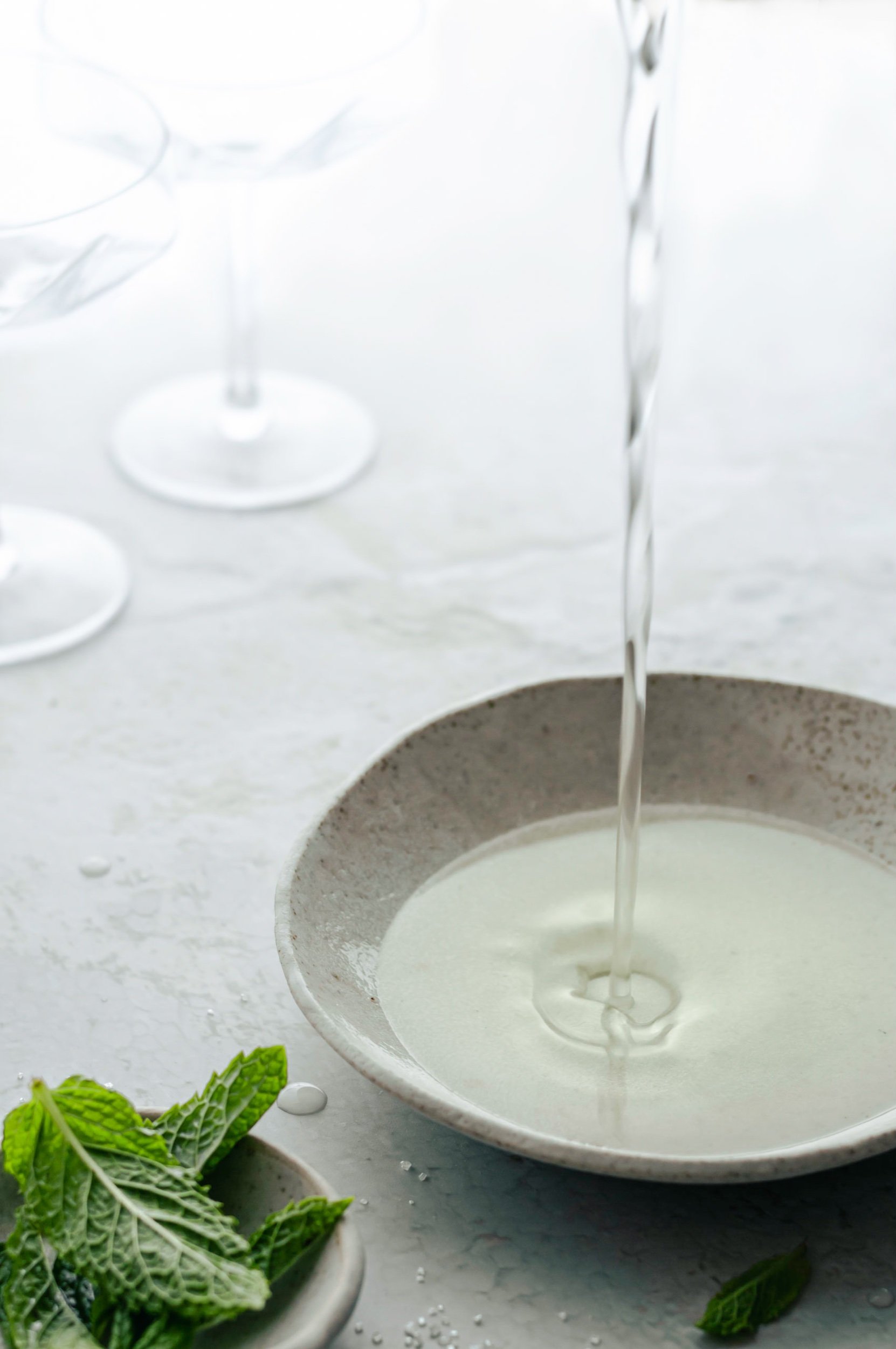hendricks mint gimlet editorial collection featuring fresh mint sugar syrup being poured into a ceramic bowl with empty cocktail glasses in the background and fresh mint leaves in the foregournd