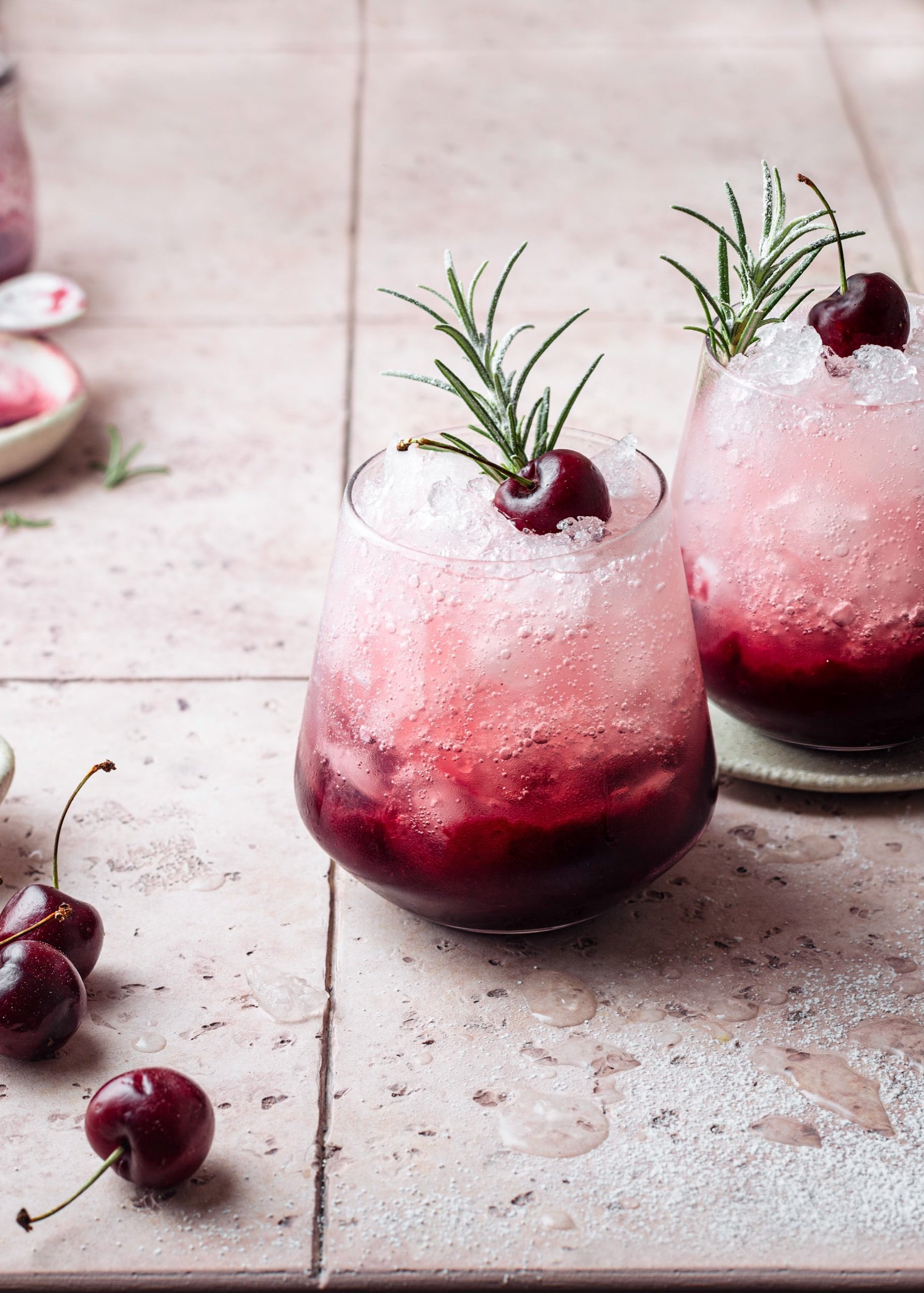 christmas cocktails cherry coolers layered with cherry syrup ice and soda water with cherry and rosemary garnish on pale pink tiled background