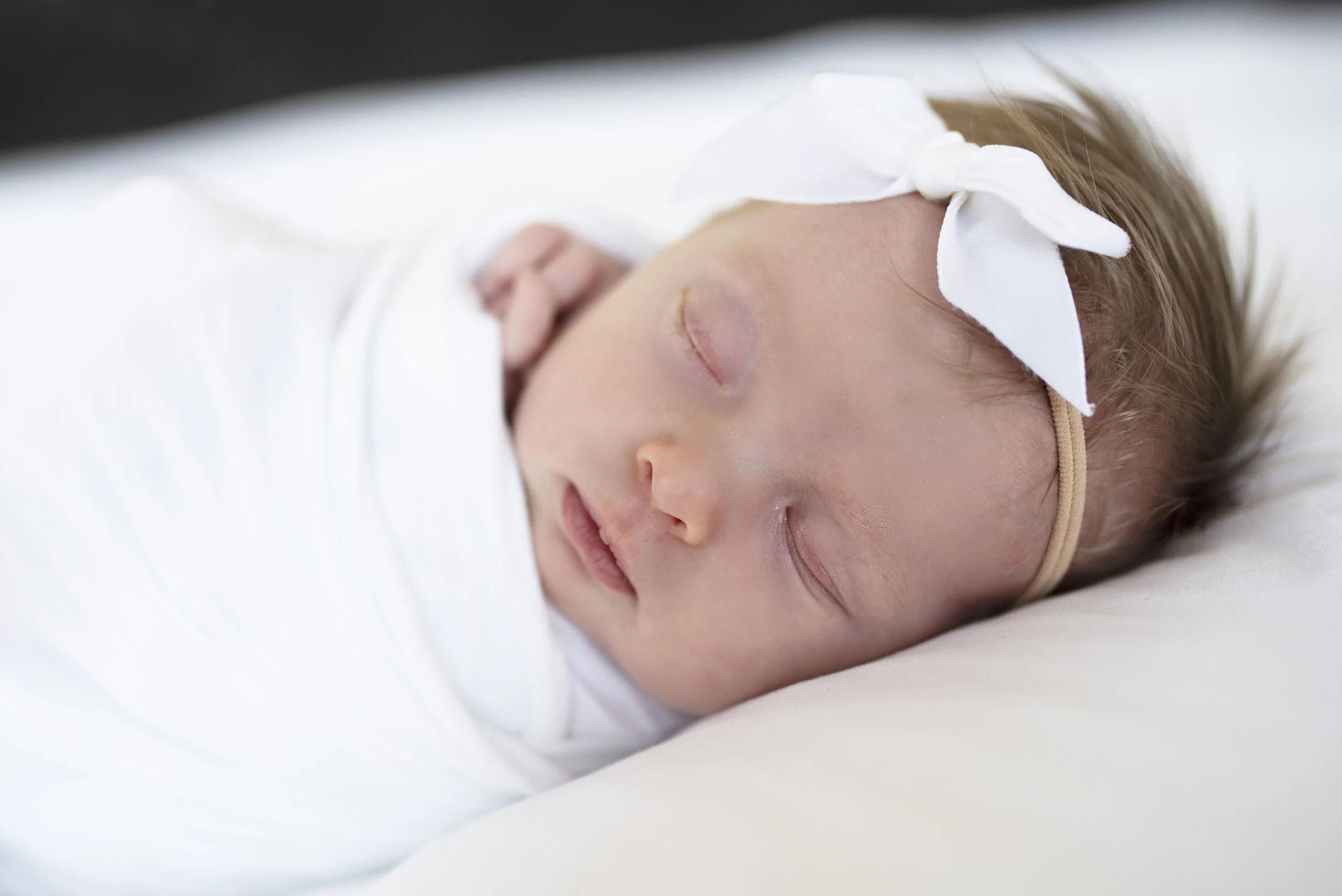 Close-up of a sleeping baby girl with a white bow headband, lying on a white pillow.