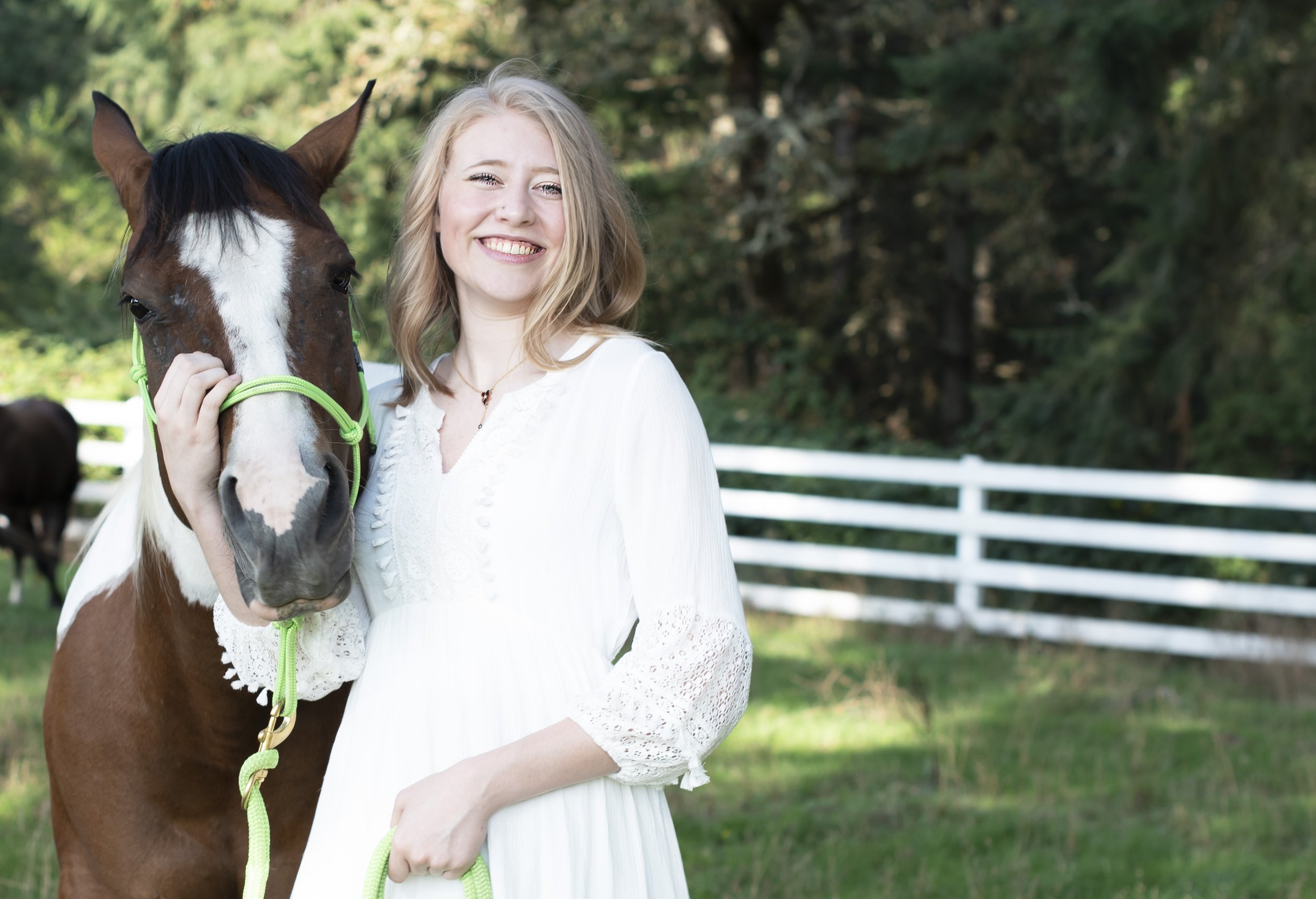 A young woman with blonde hair in a white dress smiling and holding a brown and white horse with a green halter in a grassy field with trees and a white fence in the background.