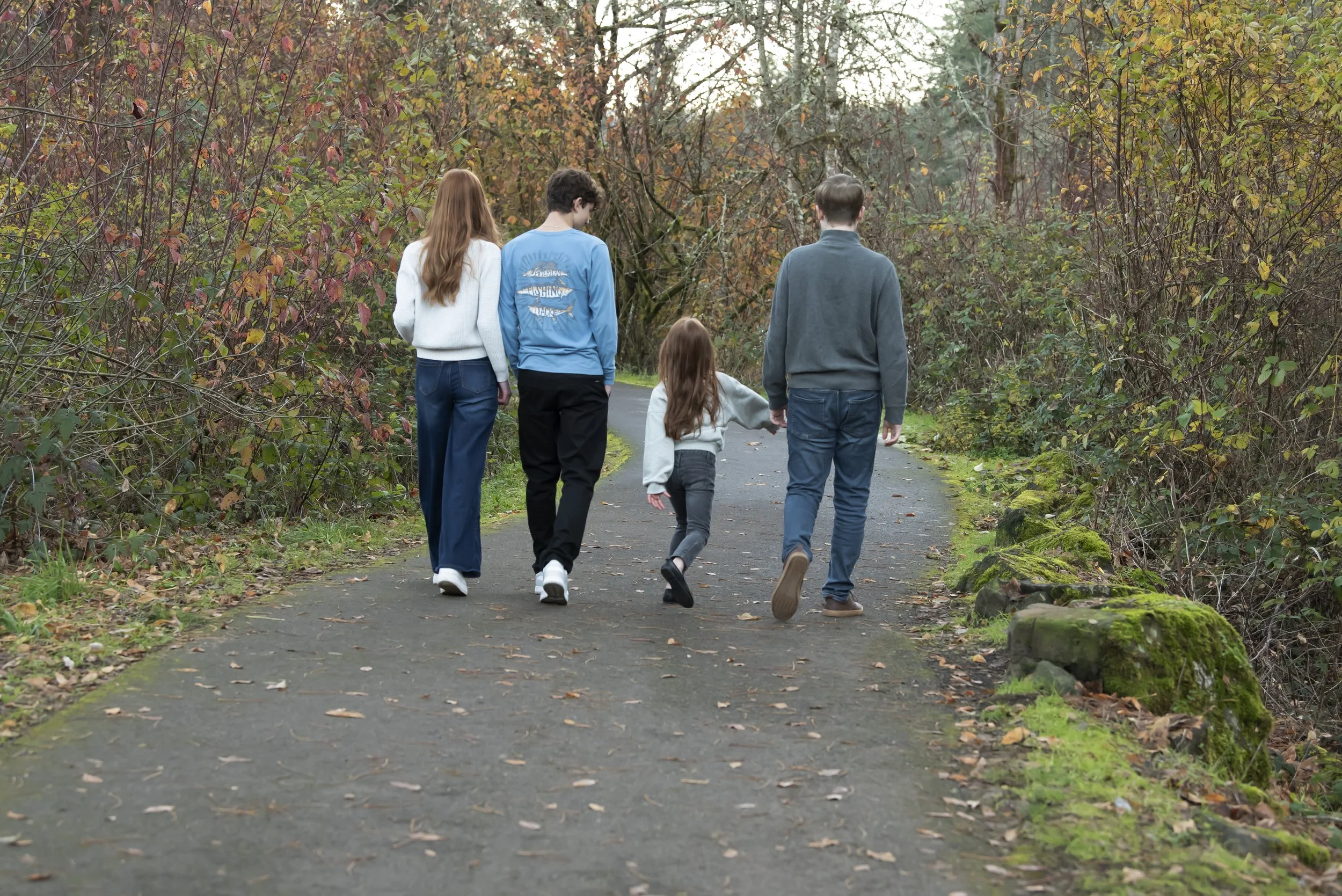 A family of five walking down a paved path through a wooded park with fall foliage.