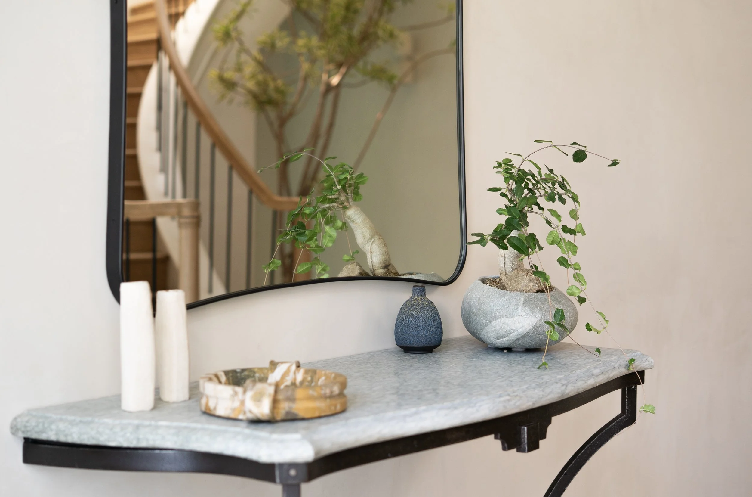 A decorative plant in a large stone pot, reflected in a wall mirror above a marble tabletop with candles and shells, in a modern interior.