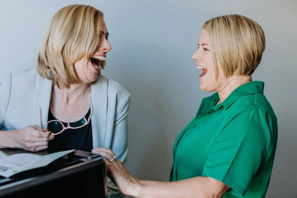 Two women laughing and smiling at each other, one holding glasses, in a friendly conversation.