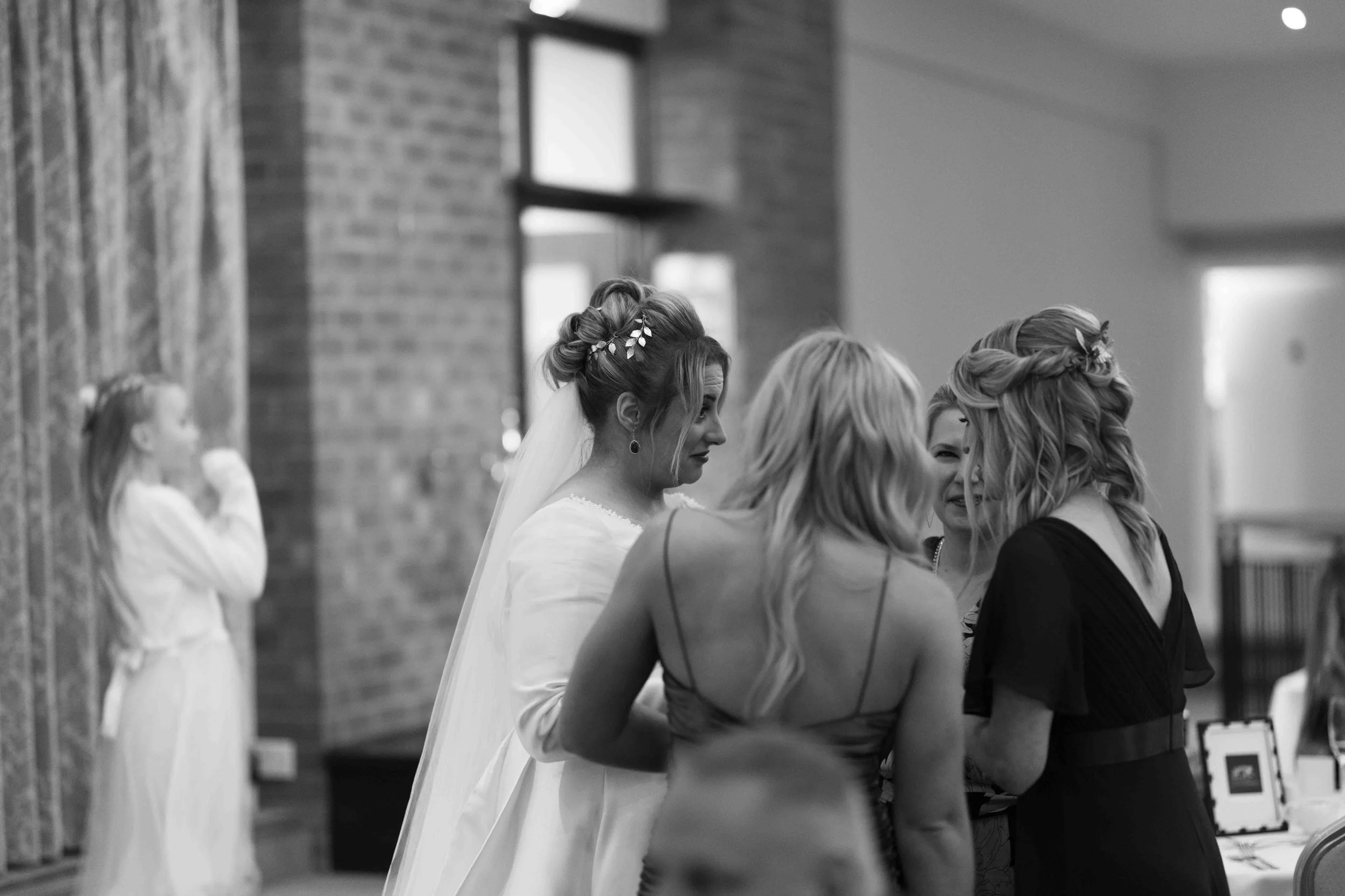 A black and white photograph of a bride in a wedding dress and veil, talking with three women at a wedding reception or gathering. A young girl in a light dress stands in the background near a brick wall.