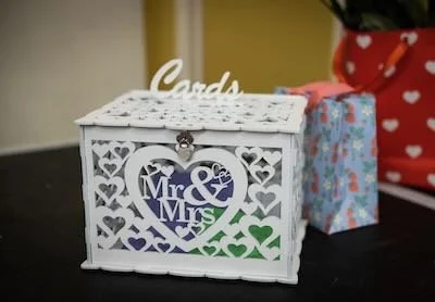 A white decorative box labeled 'Cards' with a heart-shaped cutout and 'Mr & Mrs' inside, next to wrapped gifts.