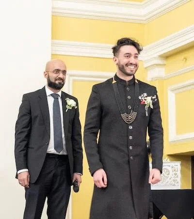 Two men in formal attire, one in a black suit and the other in a traditional black sherwani with jewelry, standing in a yellow-walled room with white trim, smiling.