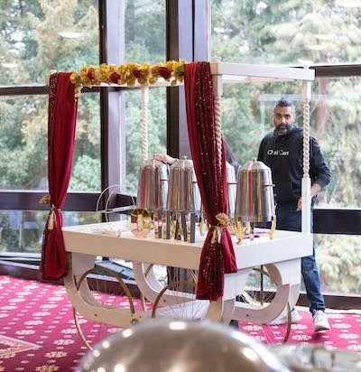 A man standing next to a decorated cart with metal containers and red curtains, in front of large windows with trees outside.
