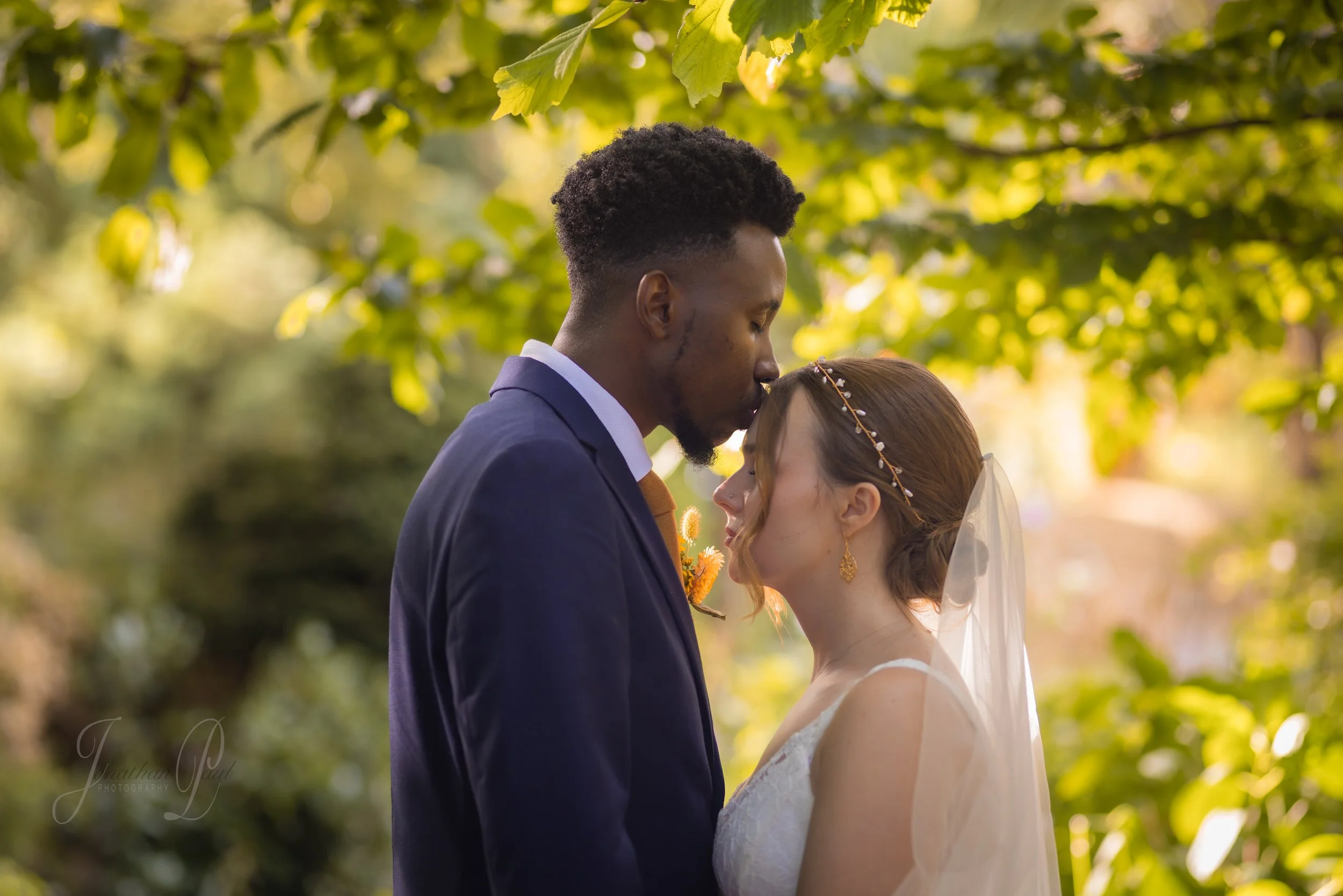 A bride and groom sharing an intimate moment outdoors, with the groom kissing the bride on her forehead against a background of green trees and sunlight.