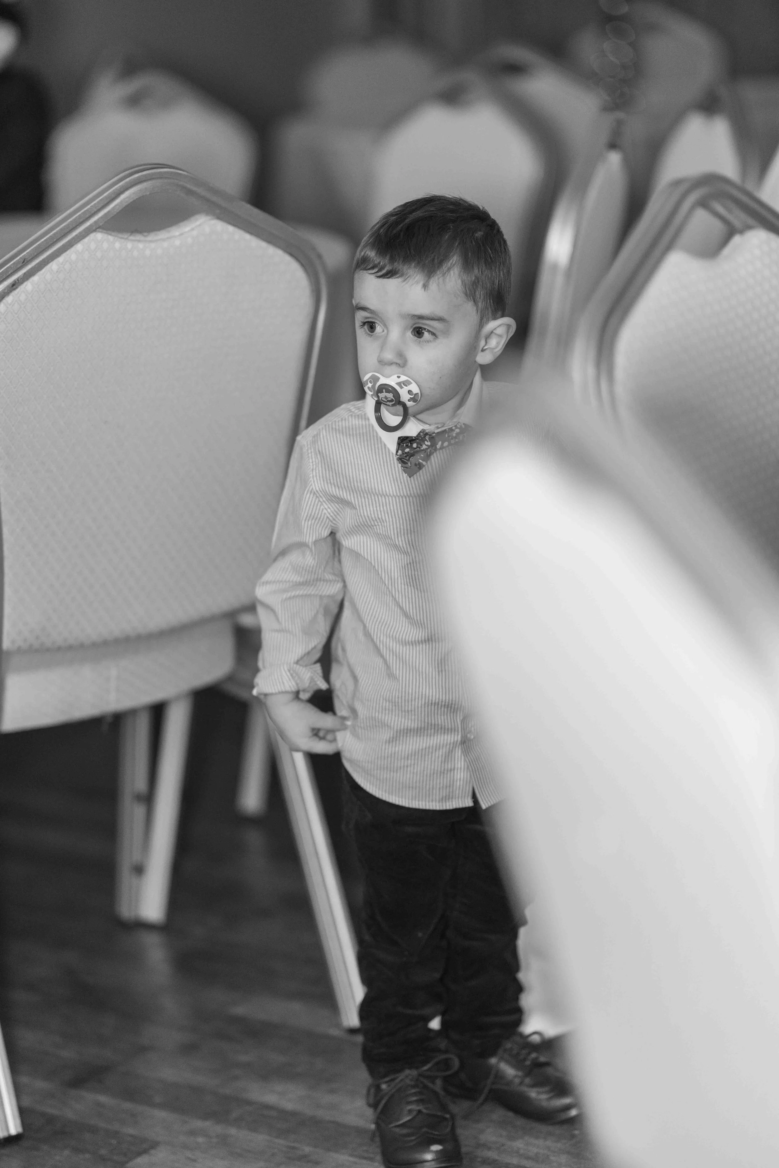 A young boy with a pacifier in his mouth, wearing a striped shirt and dark pants, standing between chairs in a room, looking to the side.