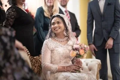 Young woman in a lace dress and veil holding a bouquet of flowers, surrounded by formally dressed people at a celebration.