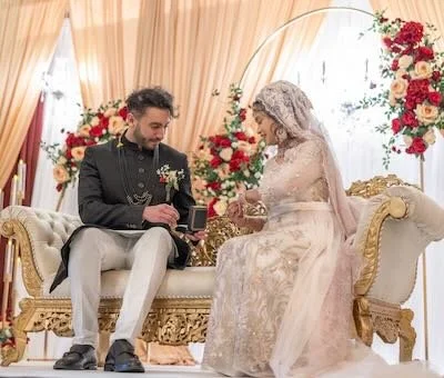 A couple sitting on a decorated sofa during a wedding ceremony, with floral arrangements and a curtain backdrop.