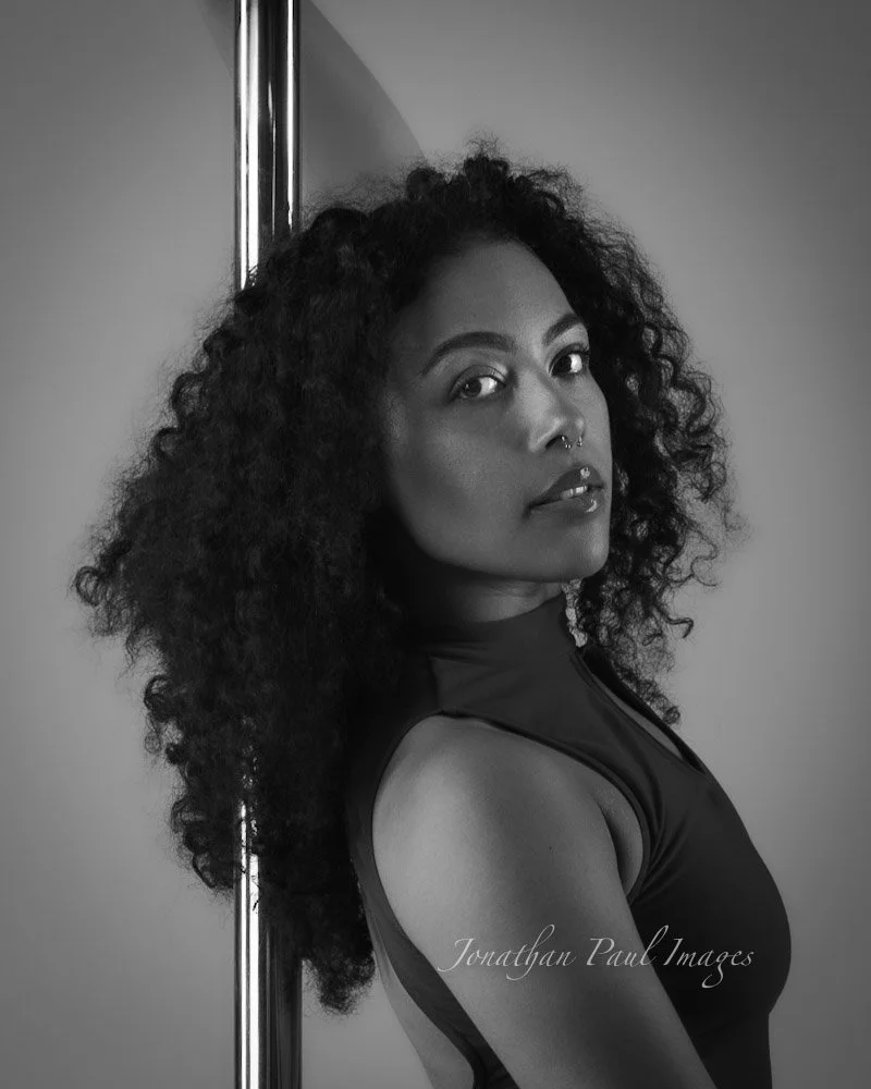 Black and white portrait of a woman with curly hair, wearing a sleeveless top, standing next to a pole, looking at the camera.