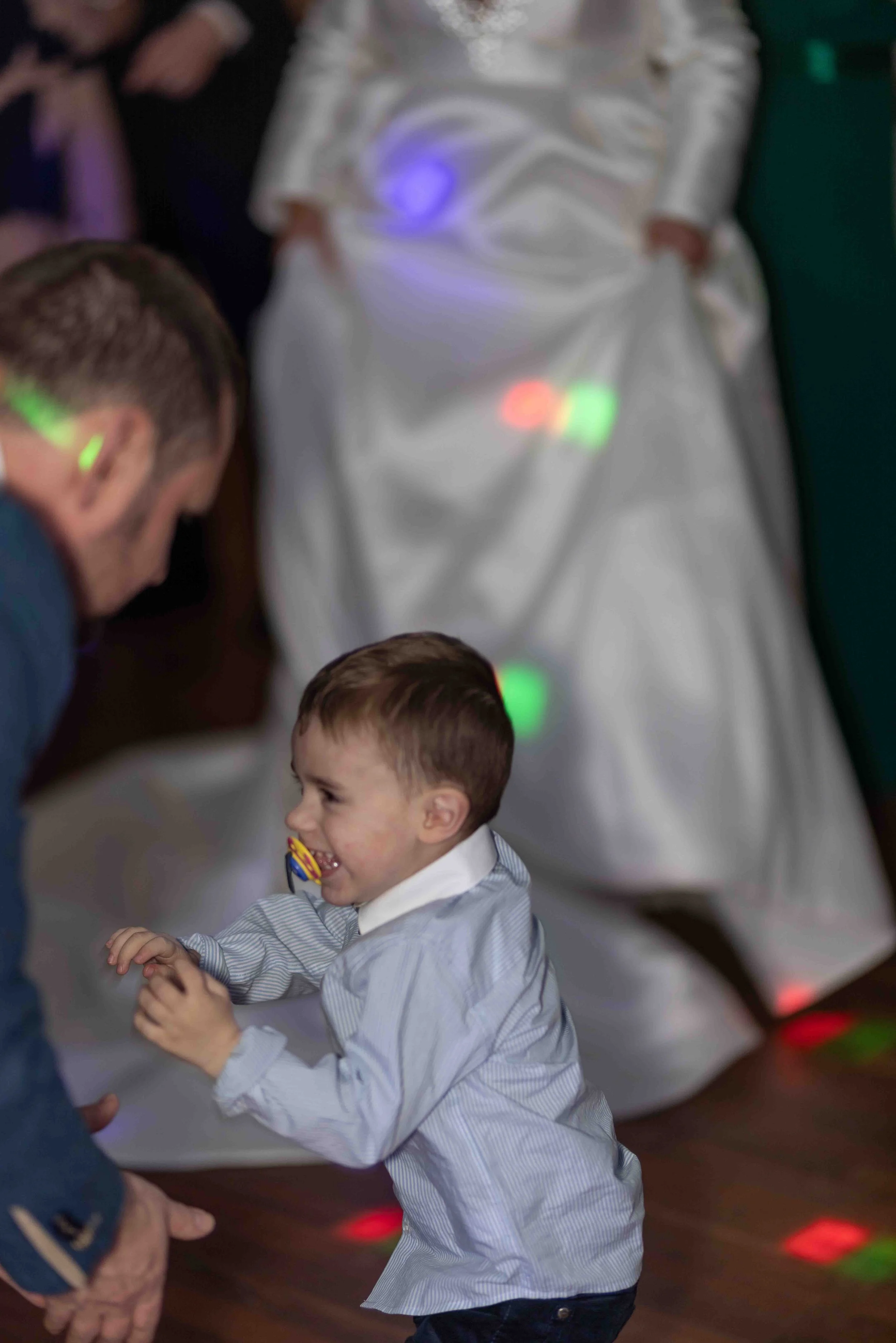 A young boy with a pacifier in his mouth laughing and dancing with an adult at a party, with a person in a white satin dress dancing in the background, colorful lights on the floor.