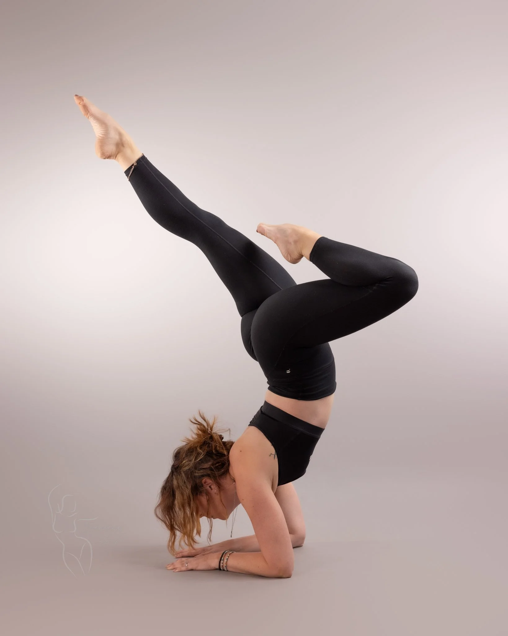 A woman in black yoga clothes performing a headstand yoga pose with her legs extending upward and bent at the knees, against a plain light background.