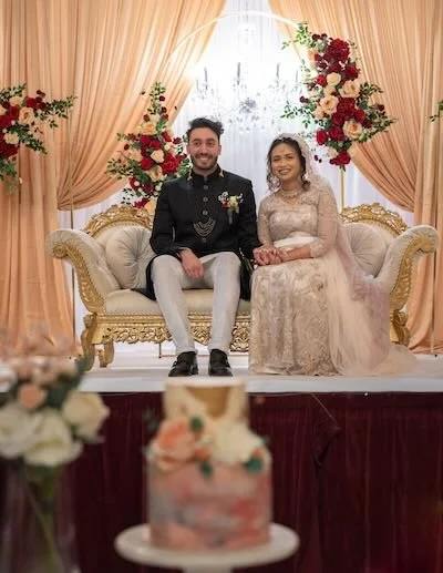 A happy couple sitting on an ornate cream-colored sofa at their wedding reception, surrounded by red and white flowers and peach-colored drapes, with a wedding cake in the foreground.