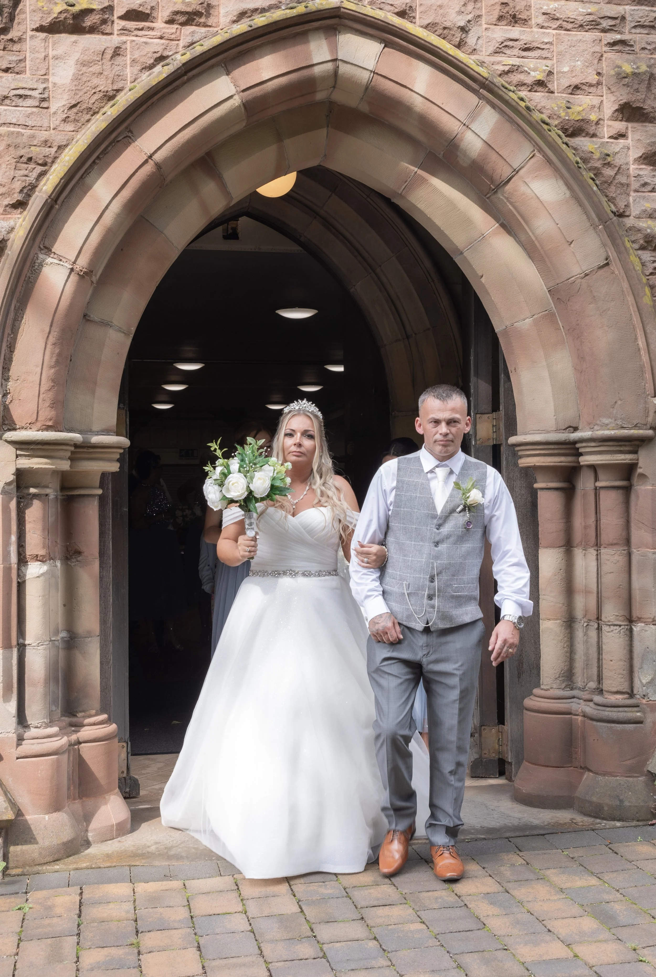 A bride in a white wedding dress holding a bouquet, walking with a groom in a gray vest and pants through a stone archway into a church.
