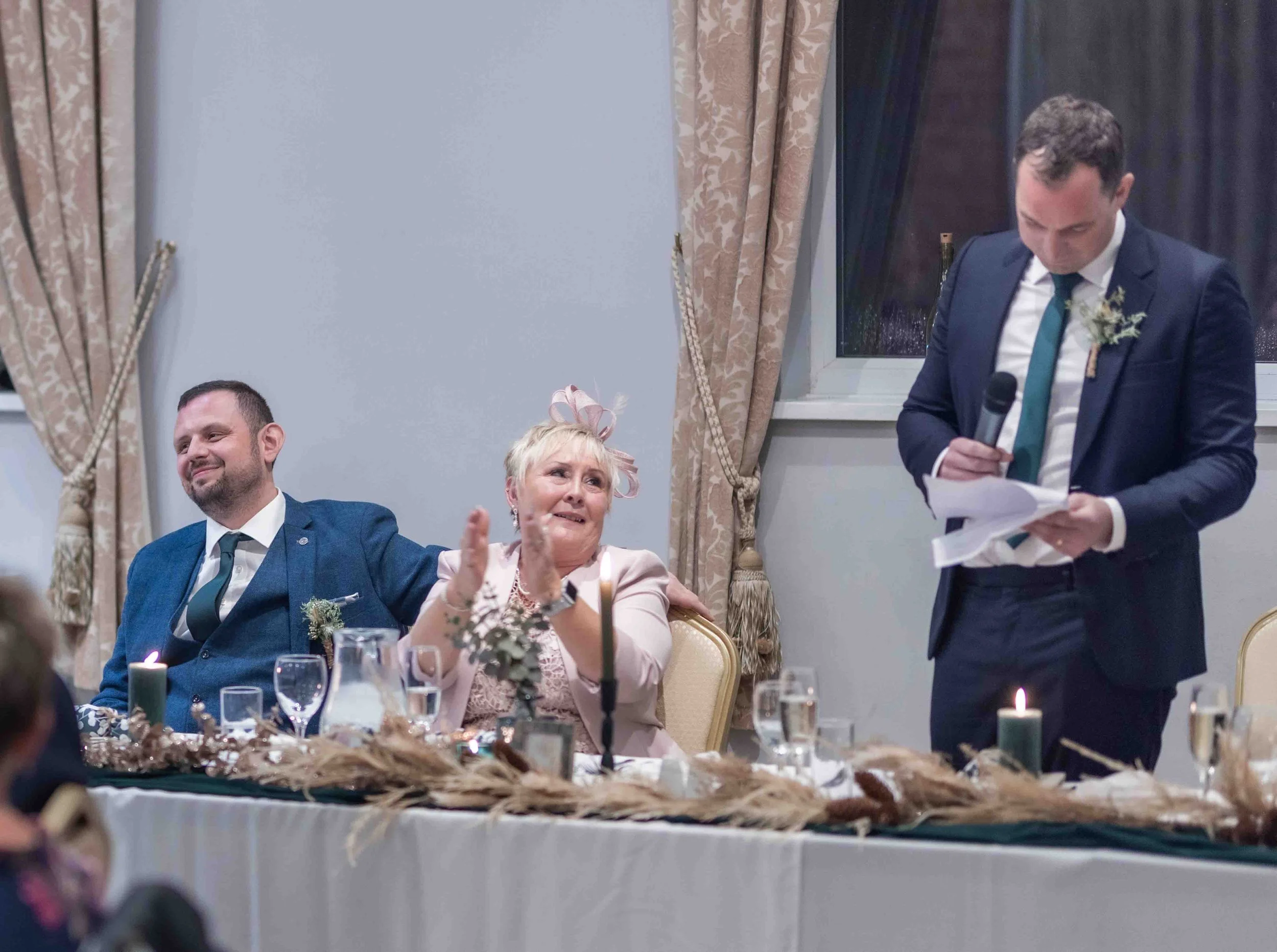 A man in a dark suit and a teal tie is giving a speech at a wedding reception, holding a microphone and reading from notes. Seated at the table are a woman in a pink dress and fascinator, and a man in a blue suit; they are clapping and smiling. The t