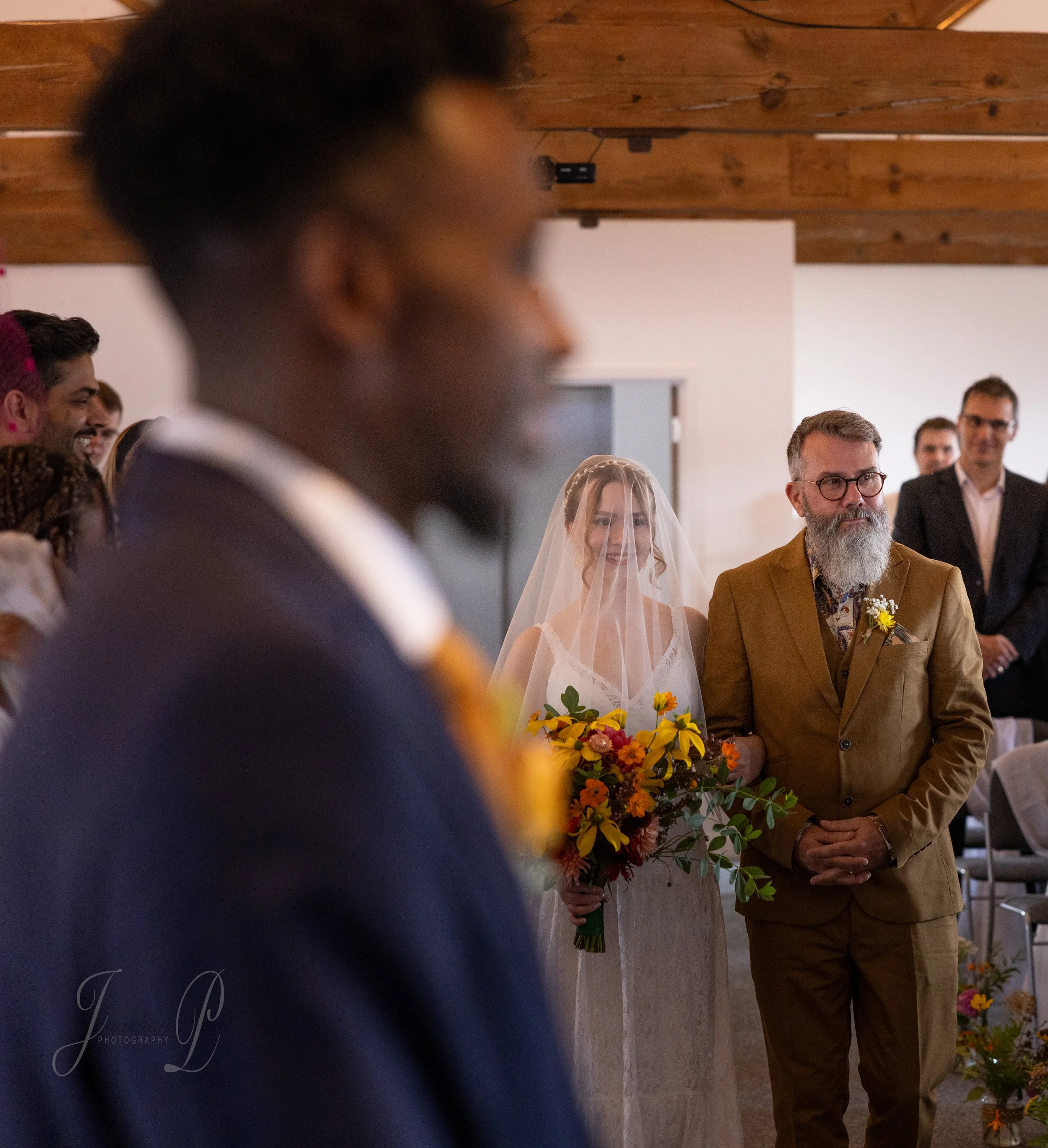 A bride with a veil holding a bouquet of flowers standing next to a man in a brown suit at a wedding ceremony. Other guests are visible in the background, smiling and attentive.