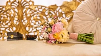 A bouquet of pink and white roses and other flowers placed on a table next to a beige folded umbrella and two small black boxes.